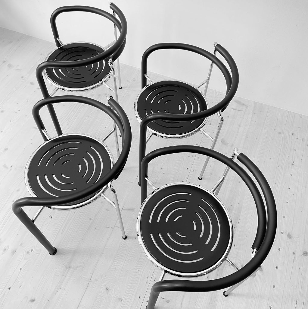 Aerial view of four modern black chairs with circular patterned seats and chrome frames, arranged closely together on a light wooden floor.