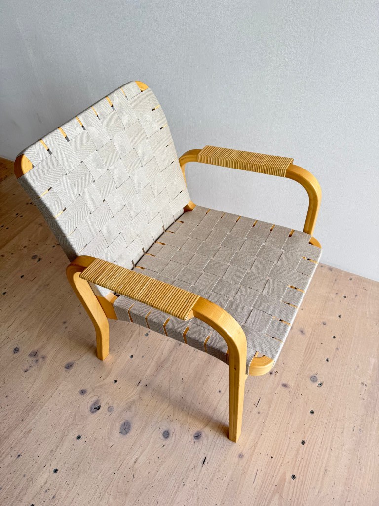 A Model 45 Dining Chair designed by Alvar Aalto, featuring a birchwood frame, cane armrests, and black linen webbing, placed on a light wooden floor.