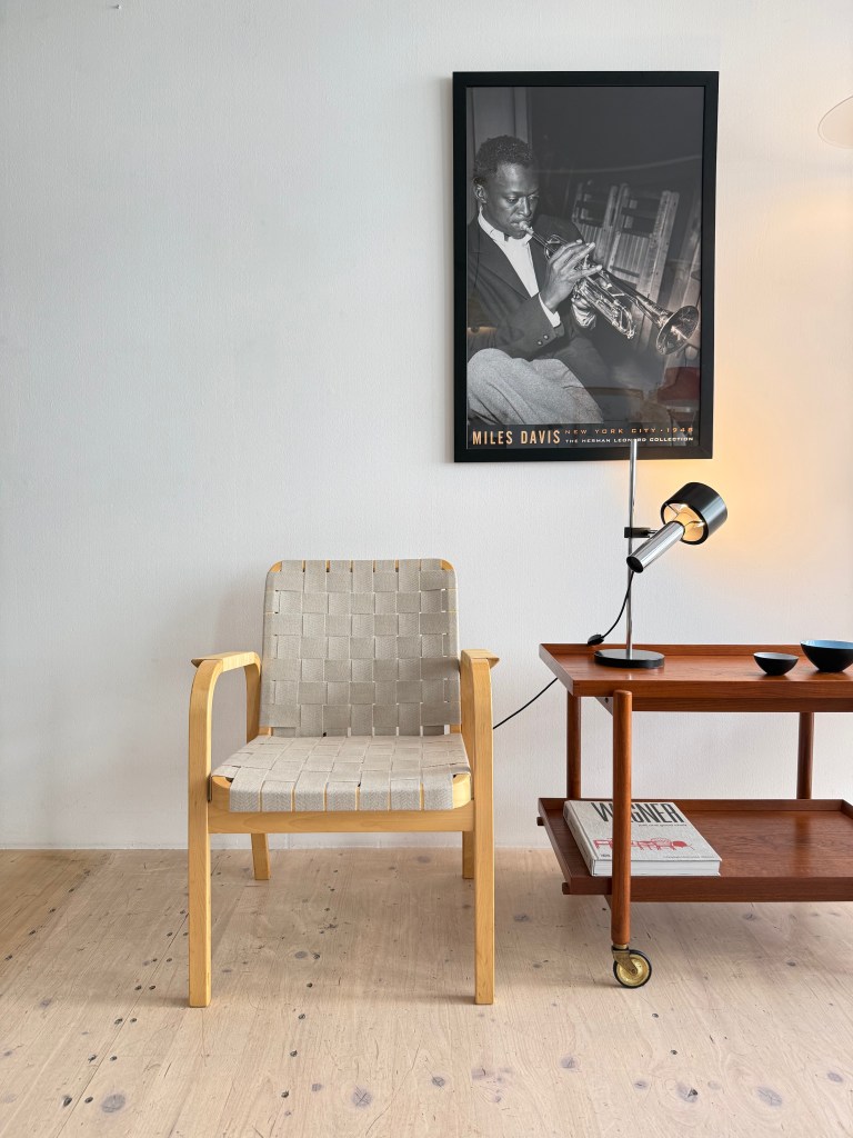 Model 45 dining chair by Alvar Aalto with a birchwood frame and cane armrests, placed next to a wooden side table with magazines and bowls, against a white wall featuring a framed black and white photo of a trumpet player.