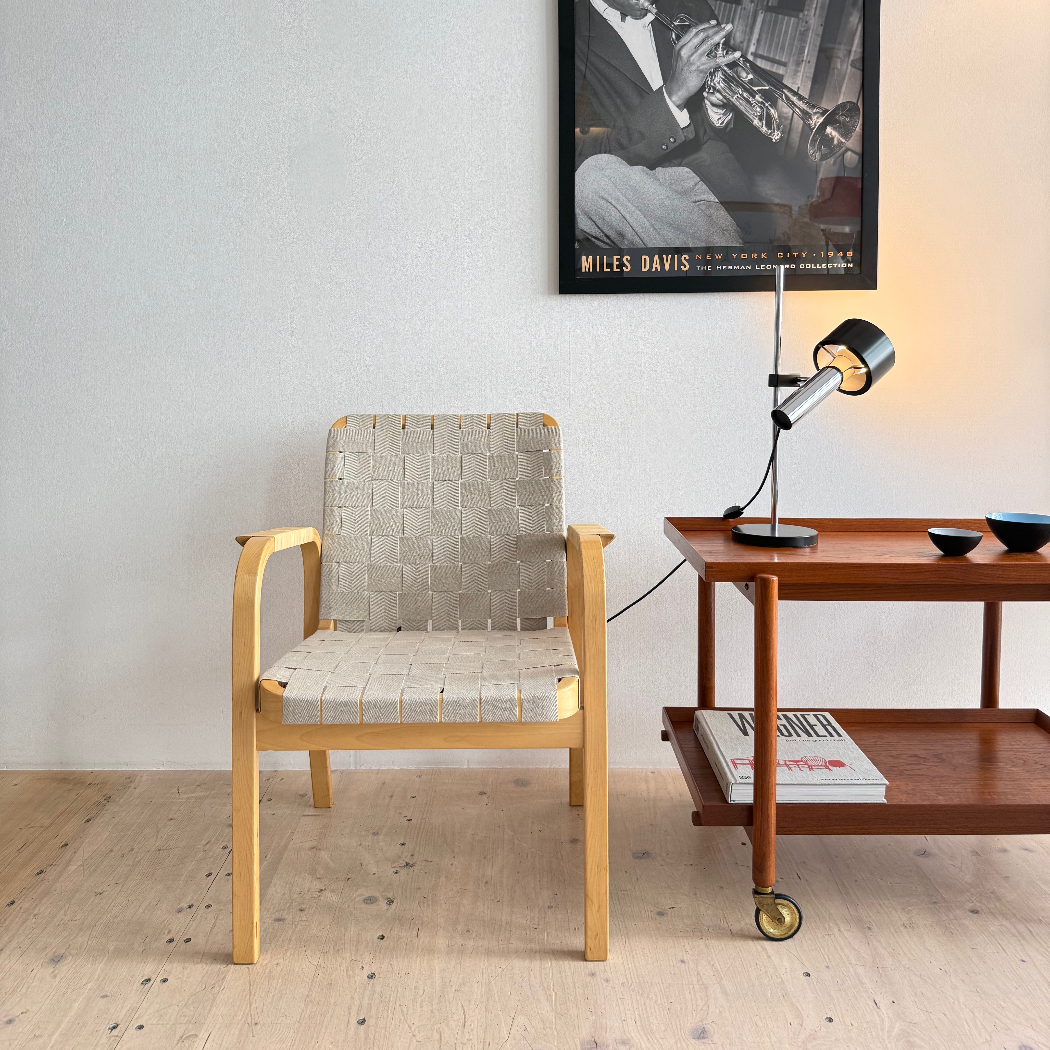 A modern chair with a woven seat, placed beside a mid-century wooden side table. The table features magazines and decorative bowls, along with a stylish lamp. A framed black and white photo of a musician, likely Miles Davis, hangs on the wall behind.