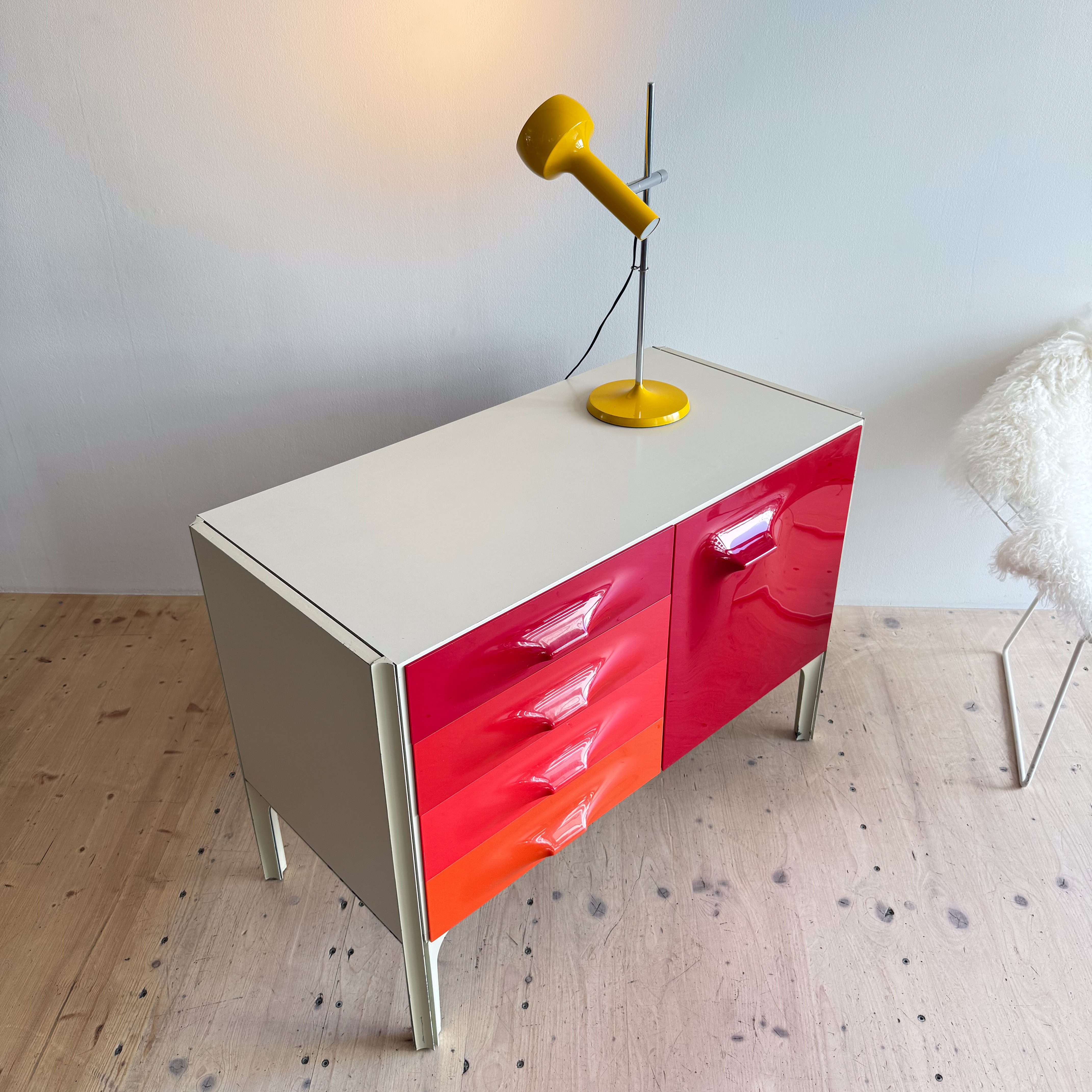 A colorful sideboard with drawers in shades of red and orange, accompanied by a yellow lamp, set against a light-colored wall.