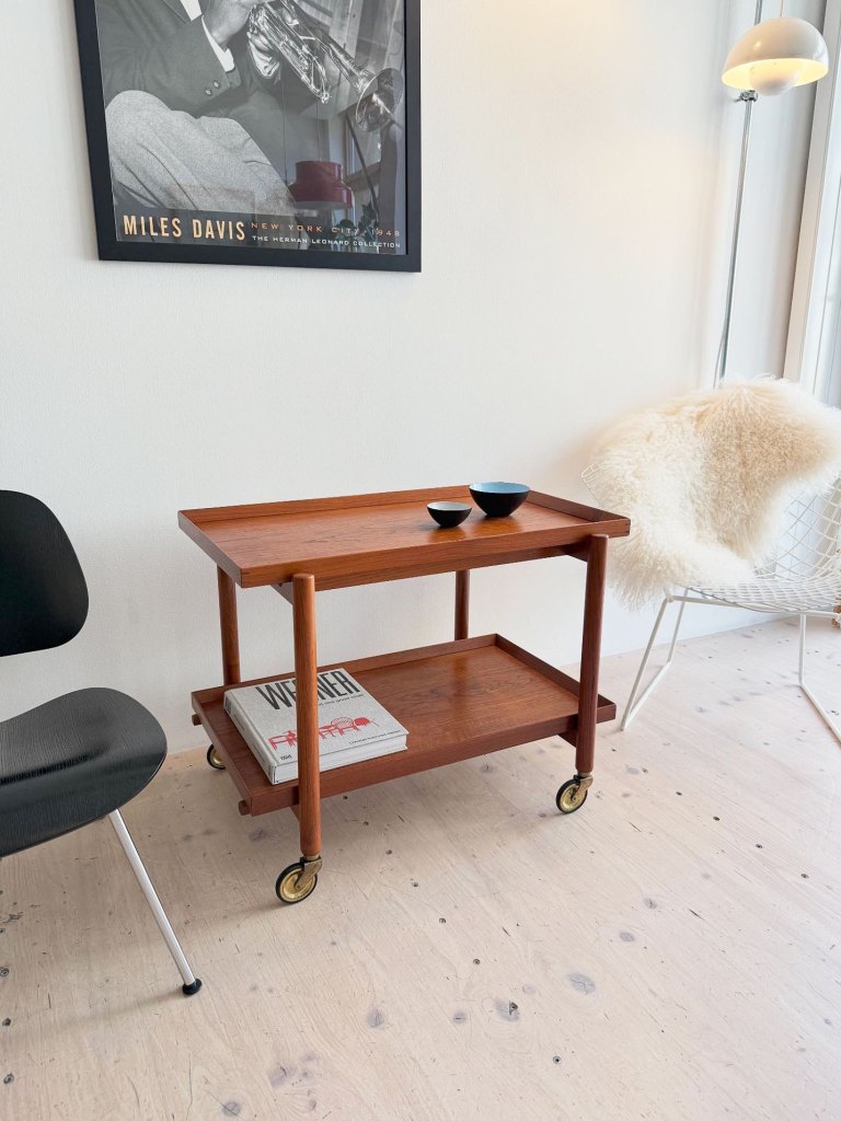 A vintage teak serving cart designed by Poul Hundevad, featuring a two-tier design, positioned in a modern room with a black chair and a decorative bowl on top.