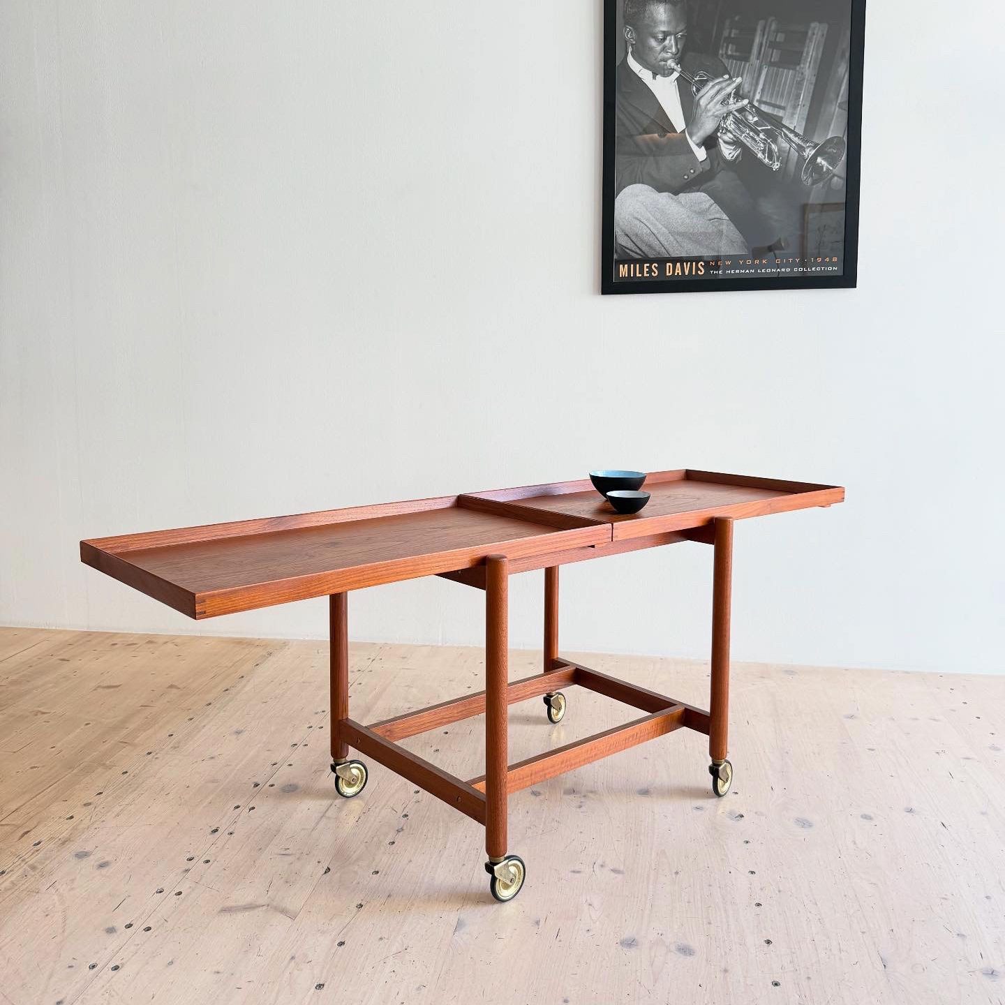 A wooden serving trolley with wheels, featuring two trays and a black bowl, set against a light-colored wall with a framed black and white poster of a musician.