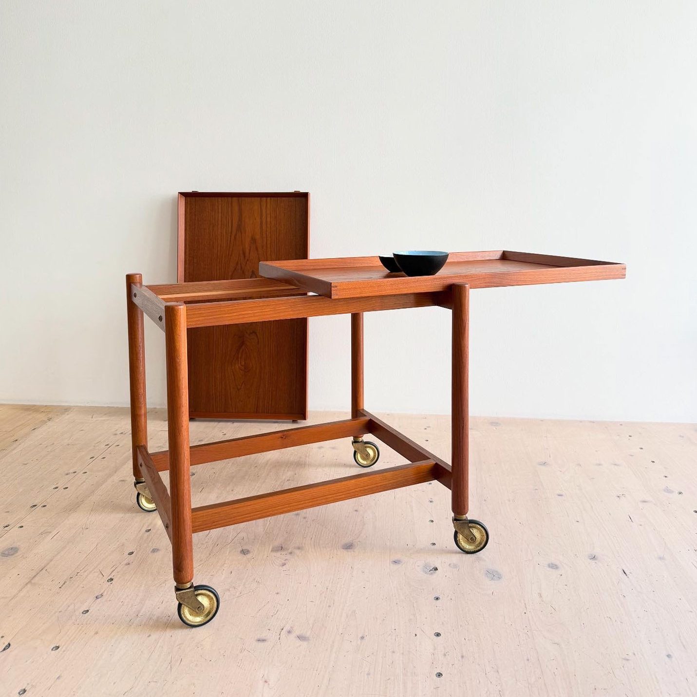 Teak serving trolley with a sliding tray and black dish on top, situated against a plain white wall.