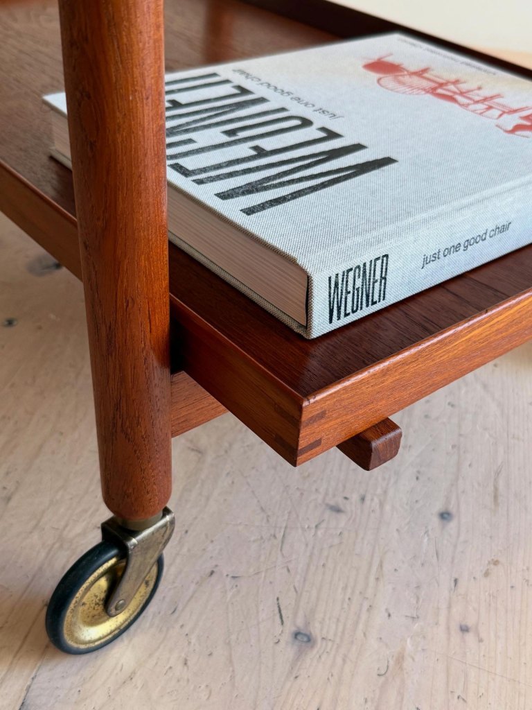 Close-up of a teak serving cart by Poul Hundevad, featuring a wooden leg, a lower tray, and a book titled 'Wegner' resting on the upper surface.