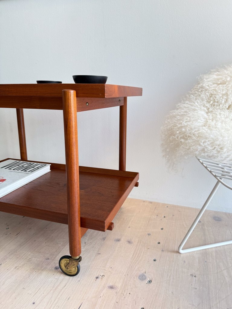Close-up of a vintage teak serving cart designed by Poul Hundevad, featuring a smooth top surface with two black bowls, a lower detachable tray, and wooden legs on wheels, set against a minimalist background.