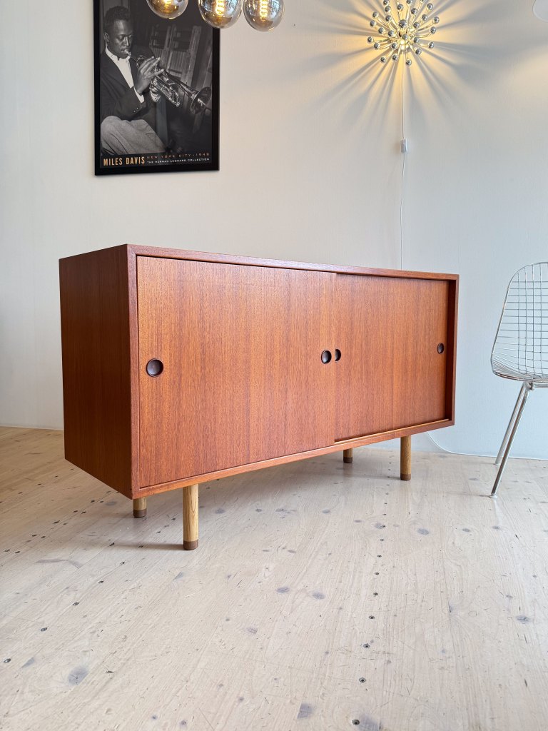 A wooden sideboard with sliding doors and round handles, sitting on wooden legs, in a well-lit room featuring a wire chair and decorative wall light, with a poster of a musician in the background.