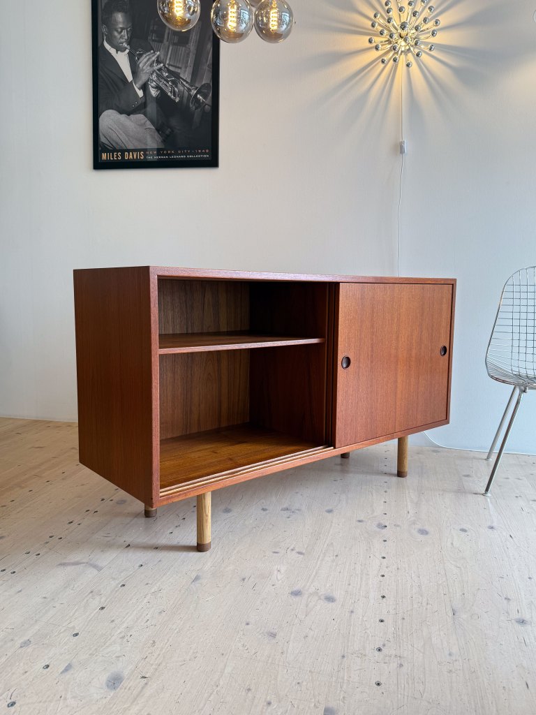 A mid-century modern wooden cabinet with two compartments, one open and one closed, sitting on wooden legs in a minimalist interior. A black and white poster of a jazz musician hangs on the wall above, alongside decorative lighting.