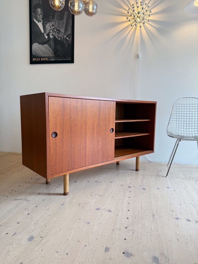 A wooden cabinet with a sliding door and open shelves, positioned on light wooden flooring, alongside a modern wire chair and decorative lighting.