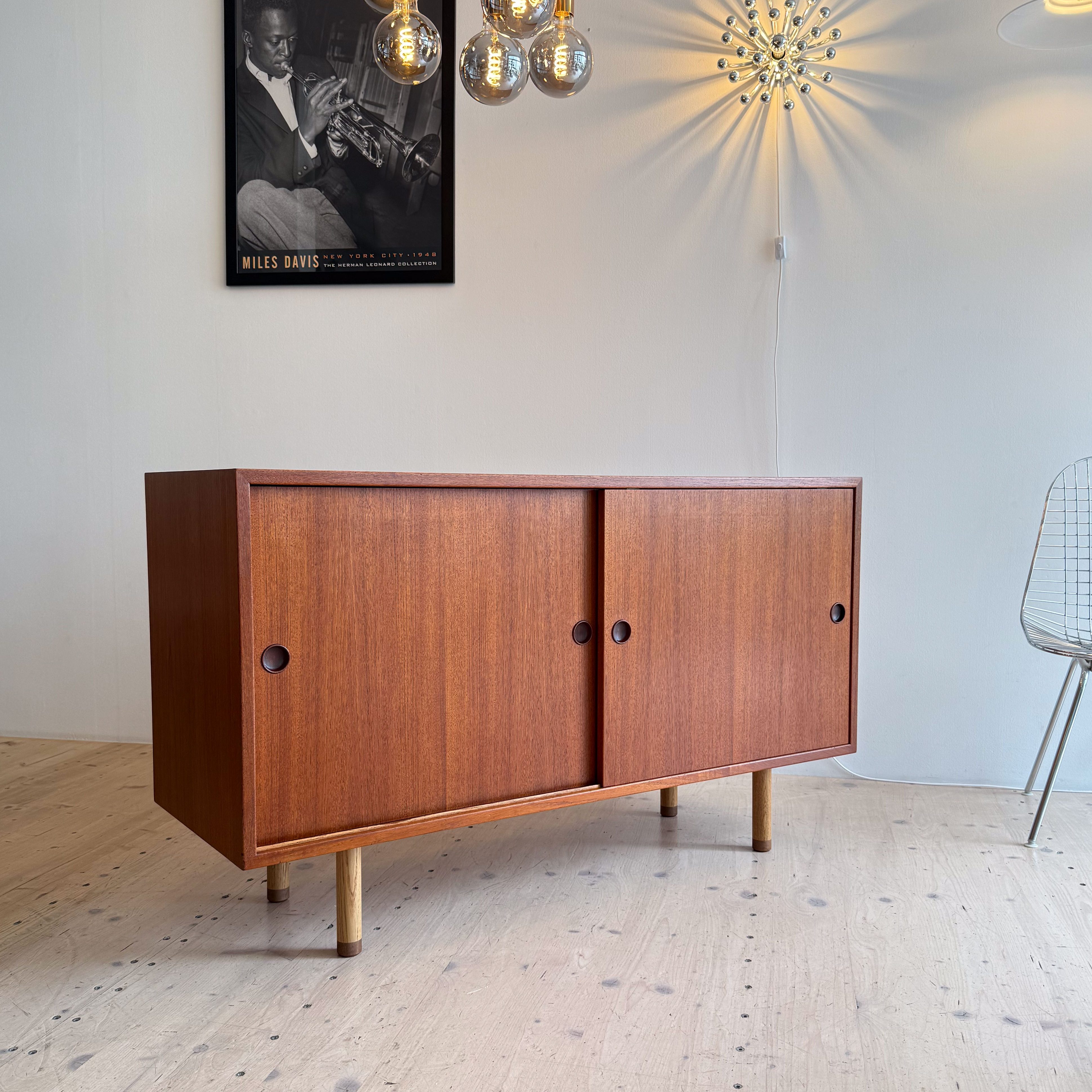 A stylish wooden sideboard with two doors, set in a modern interior featuring a decorative chandelier and a wall-mounted artwork of a musician.