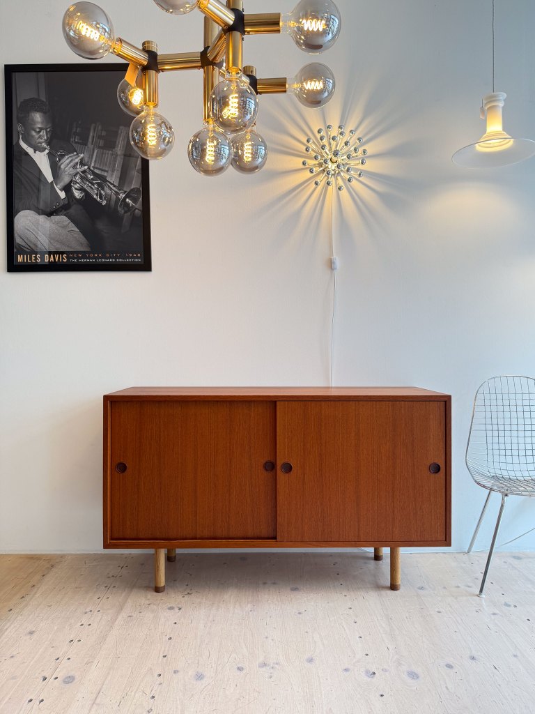 A stylish wooden cabinet with sliding doors, accompanied by a modern chandelier and decorative wall light, set against a white wall. A framed black-and-white photo of a musician is displayed above the cabinet.