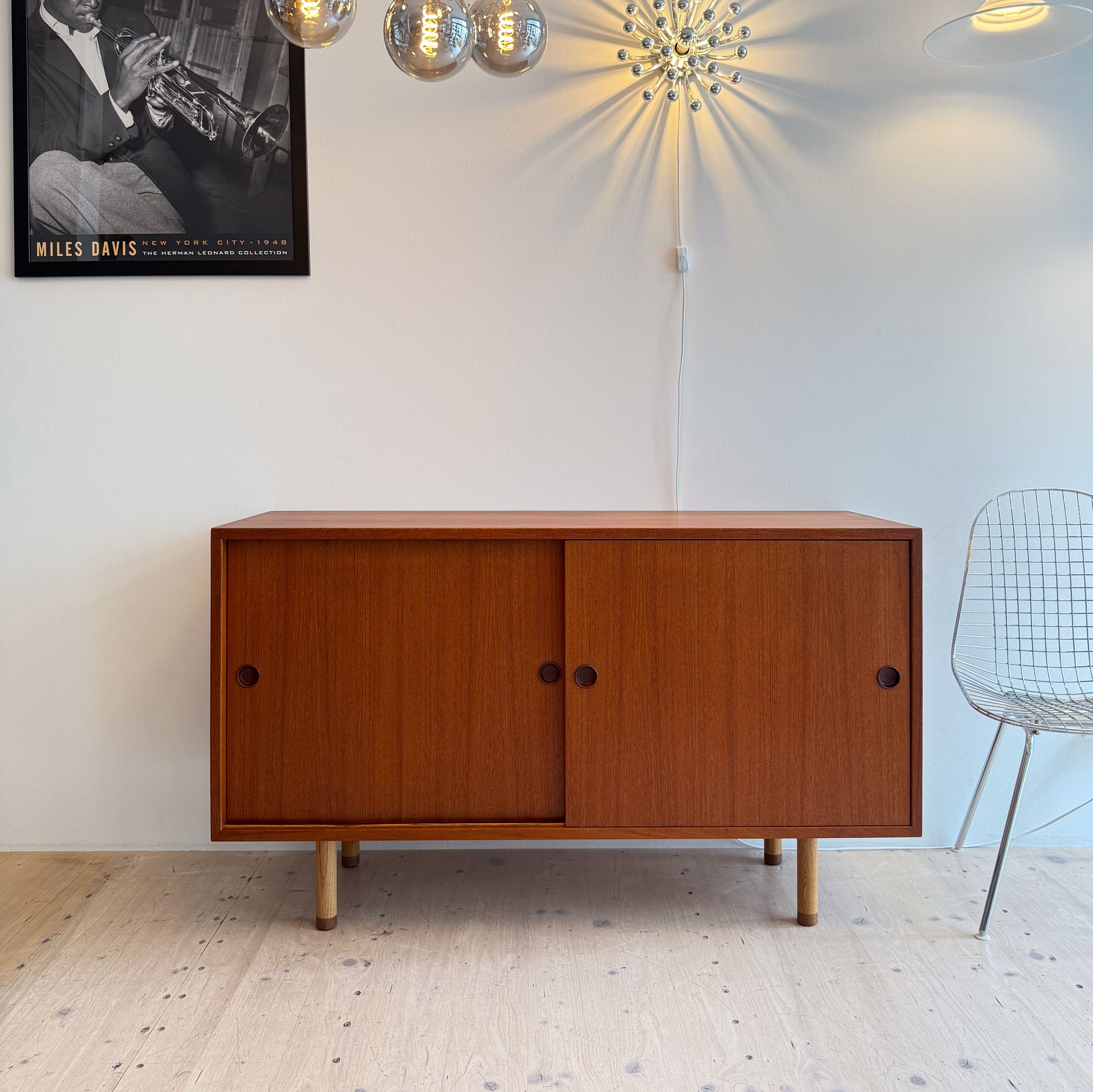 A modern teak sideboard with sliding doors and wooden legs, situated in a minimalist room featuring a stylish pendant light and an artistic wall light, accompanied by a decorative poster of a jazz musician.