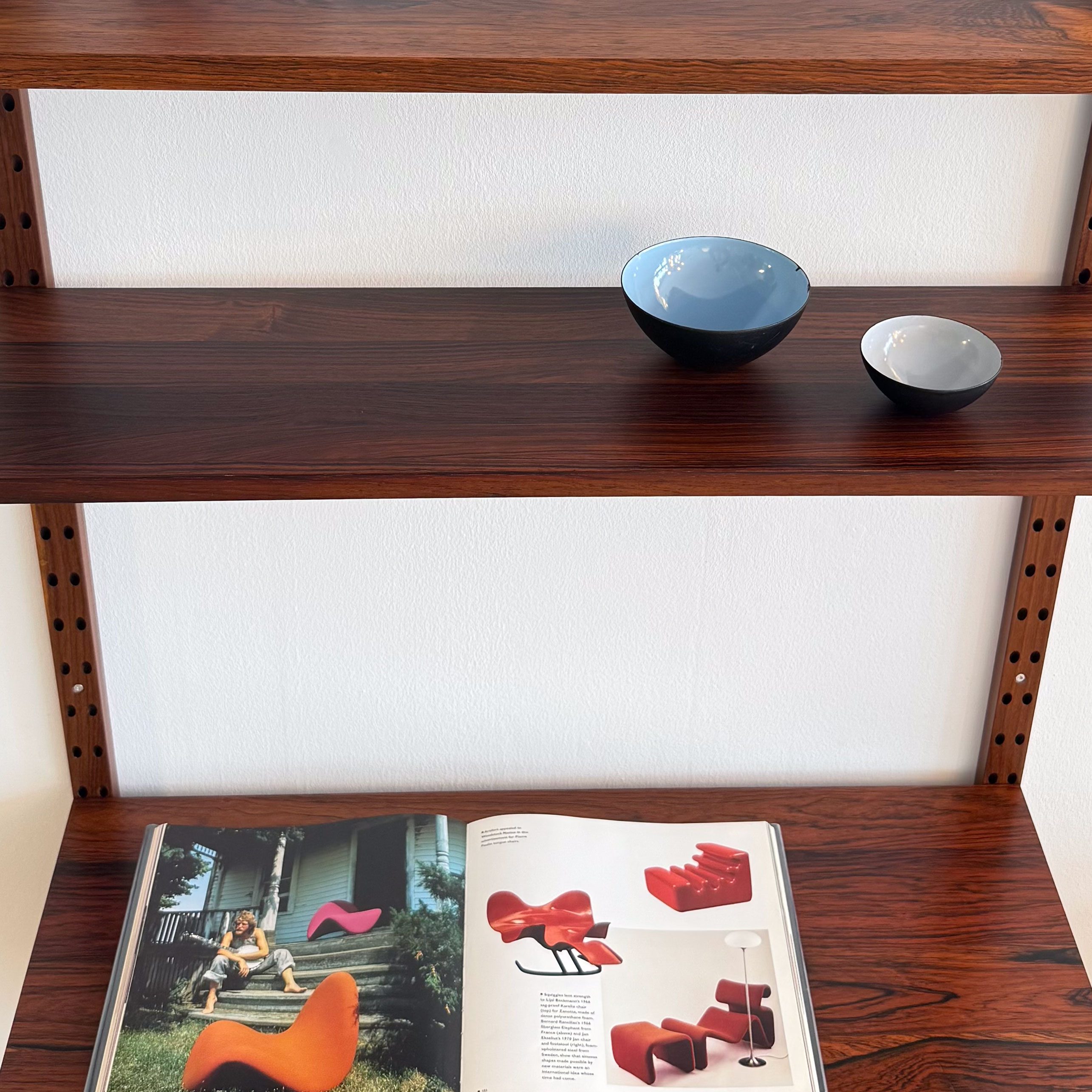 A wooden shelving unit with three shelves, featuring two decorative bowls and an open book displaying furniture designs.