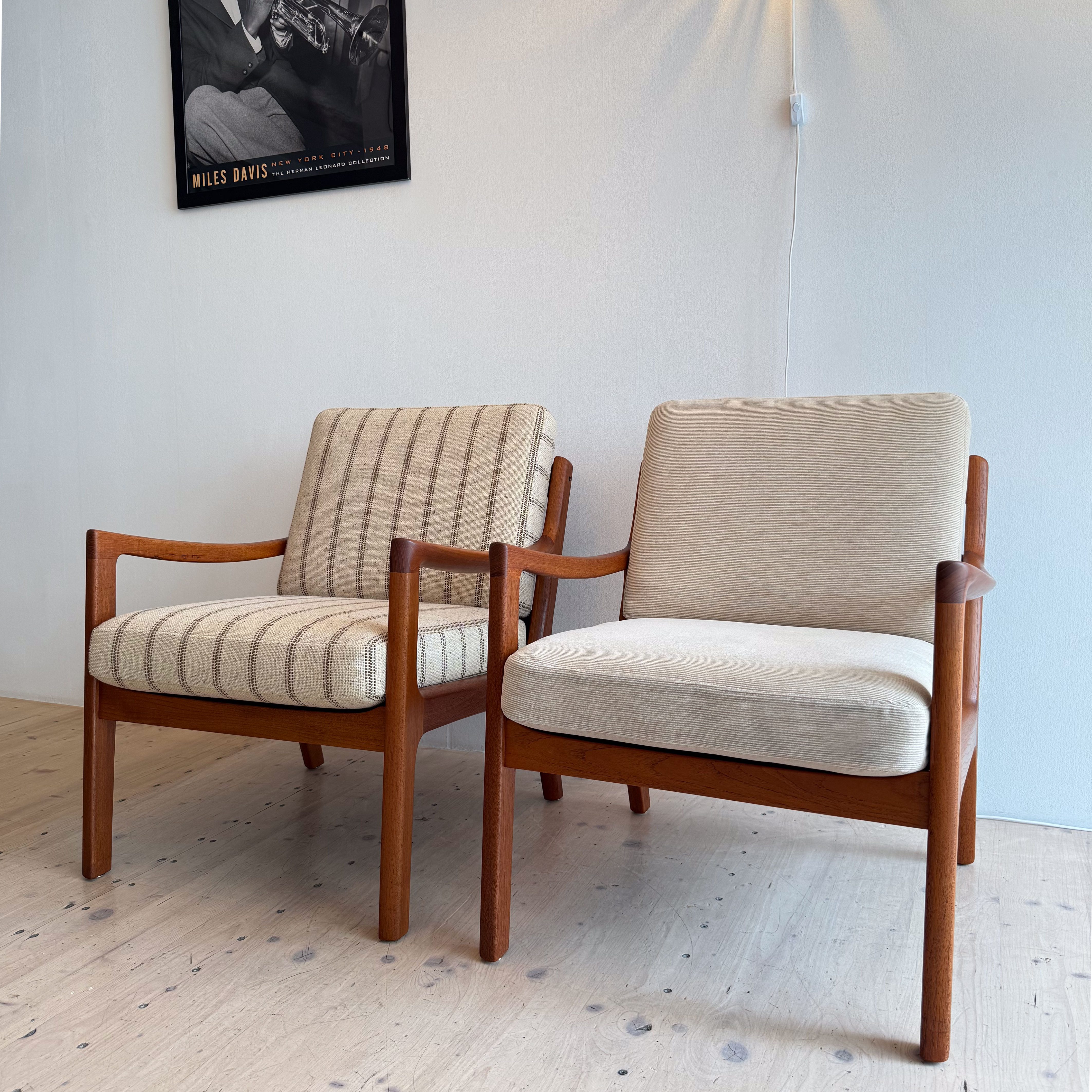 A pair of vintage lounge chairs with wooden frames and patterned upholstery, set against a light-colored wall with a pendant light and a black and white photo of a musician.