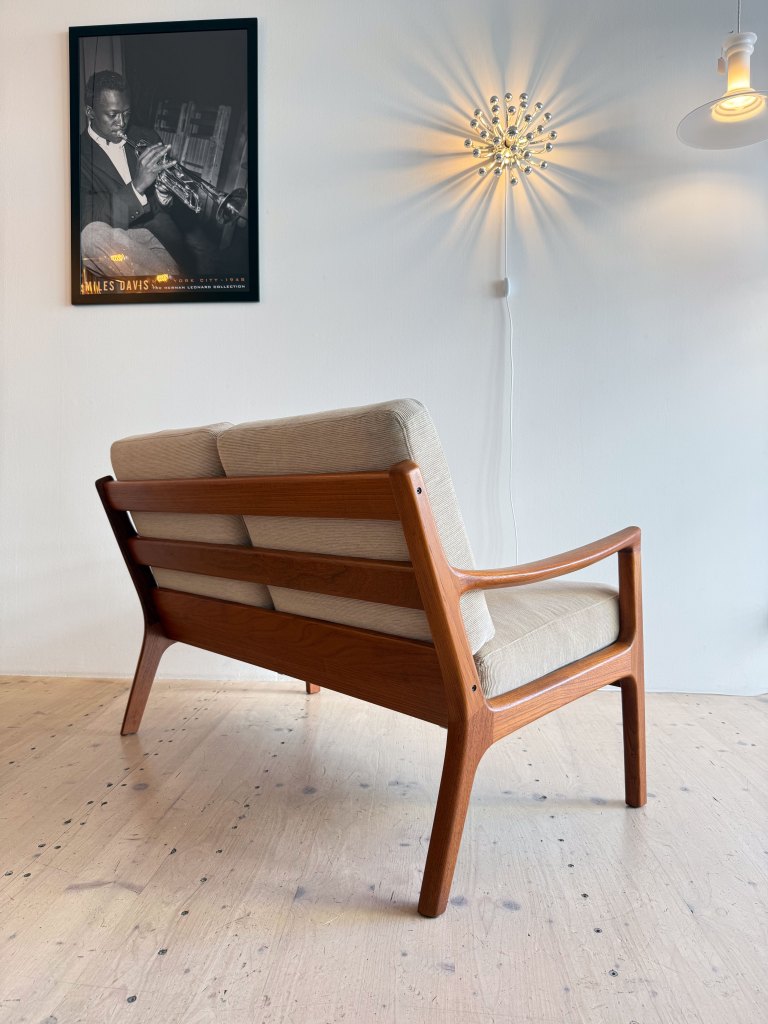A mid-century modern wooden chair with a beige cushion, positioned against a white wall. A framed black and white photo of a musician playing the trumpet hangs above, alongside a unique wall light fixture casting a starburst pattern.
