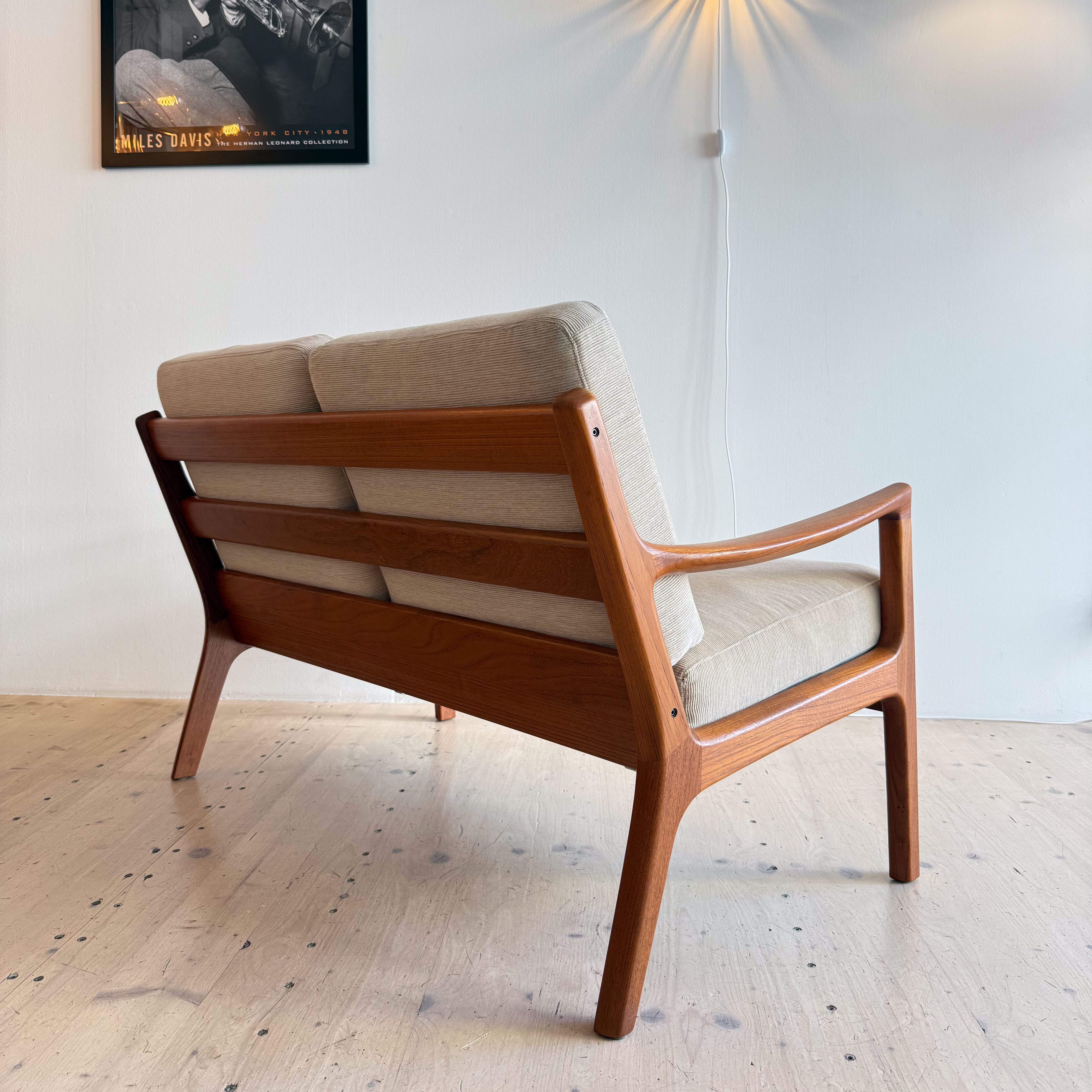 A stylish wooden lounge chair with a fabric cushion, positioned beside a trumpet player poster and a starburst lamp on a white wall.