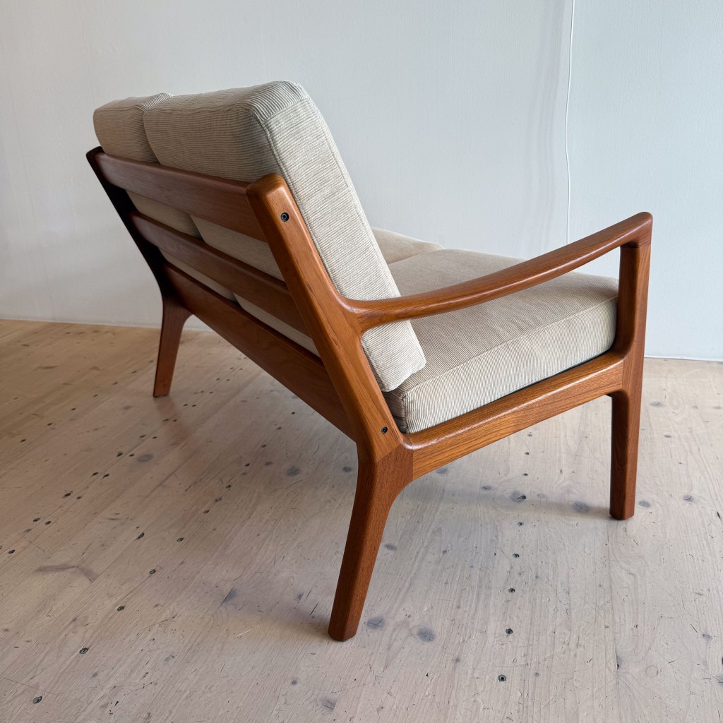 A mid-century modern wooden chair with a light fabric cushion, shown from the side and slightly angled towards the back.