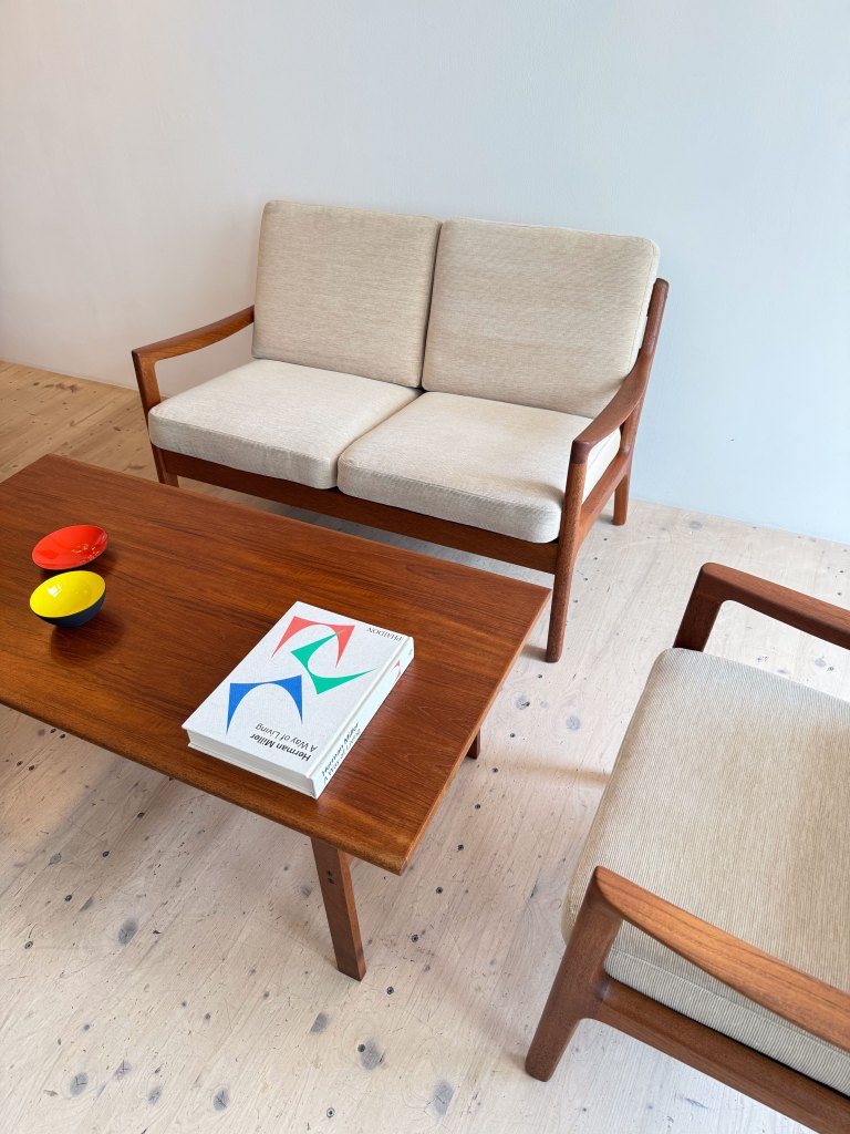 A cozy living room setup featuring a light-colored sofa and armchair with wooden frames, accompanied by a wooden coffee table displaying a book and two colorful bowls.