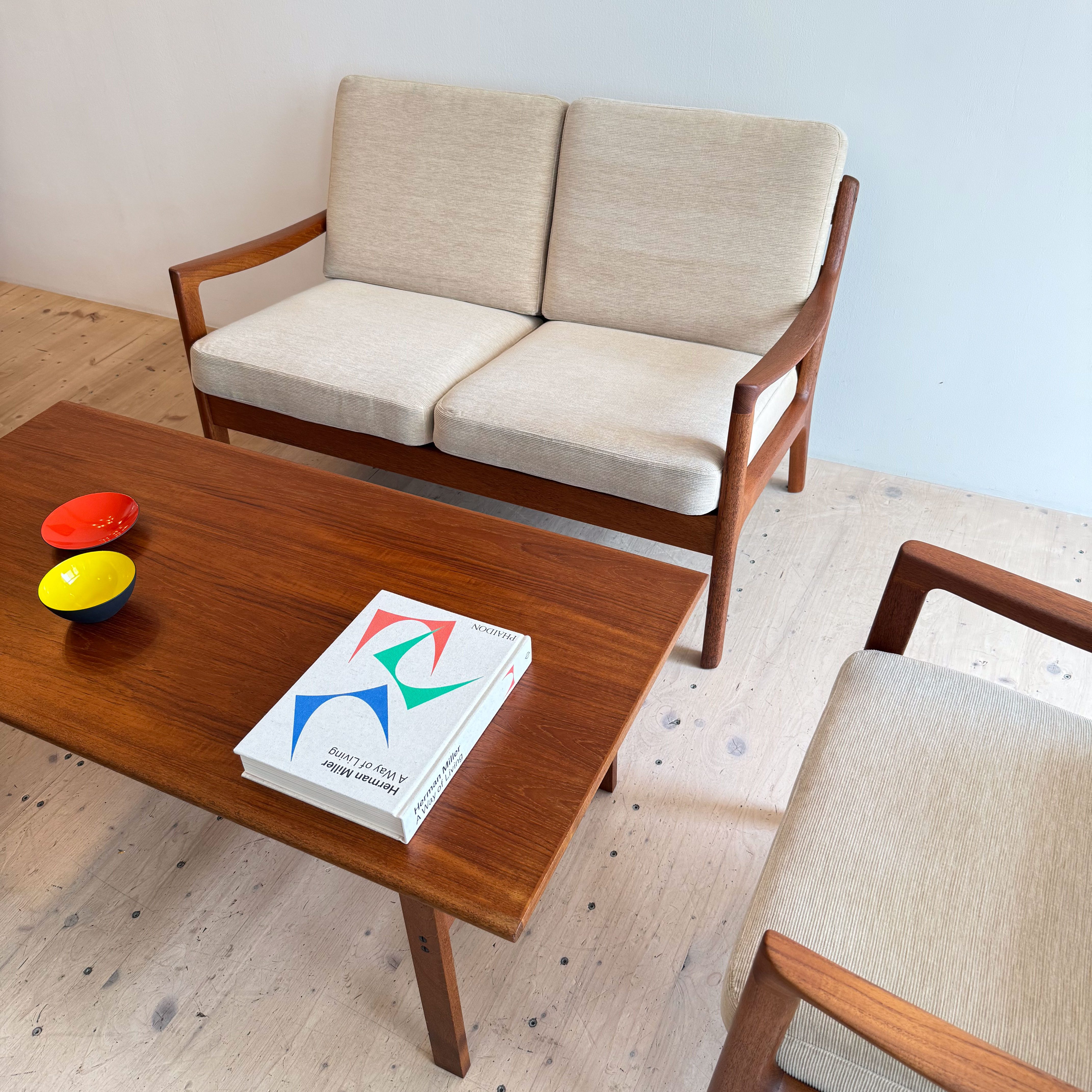 A stylish seating arrangement featuring a teak two-seater sofa with light beige cushions, accompanied by a matching armchair, set on a wooden floor with a coffee table that holds a book and colorful bowls.