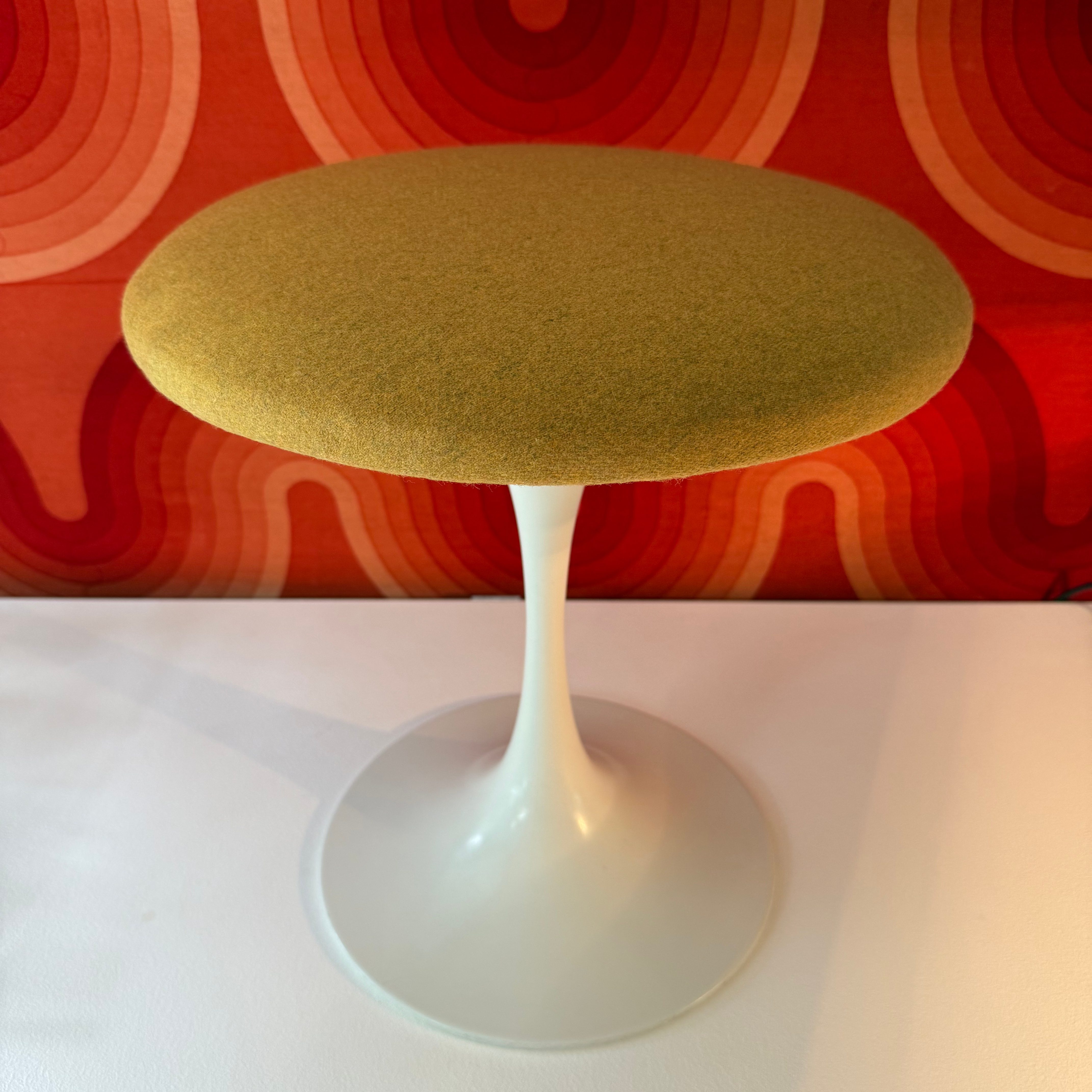 Green upholstered stool with a white base, set against a retro-style orange and red patterned background.