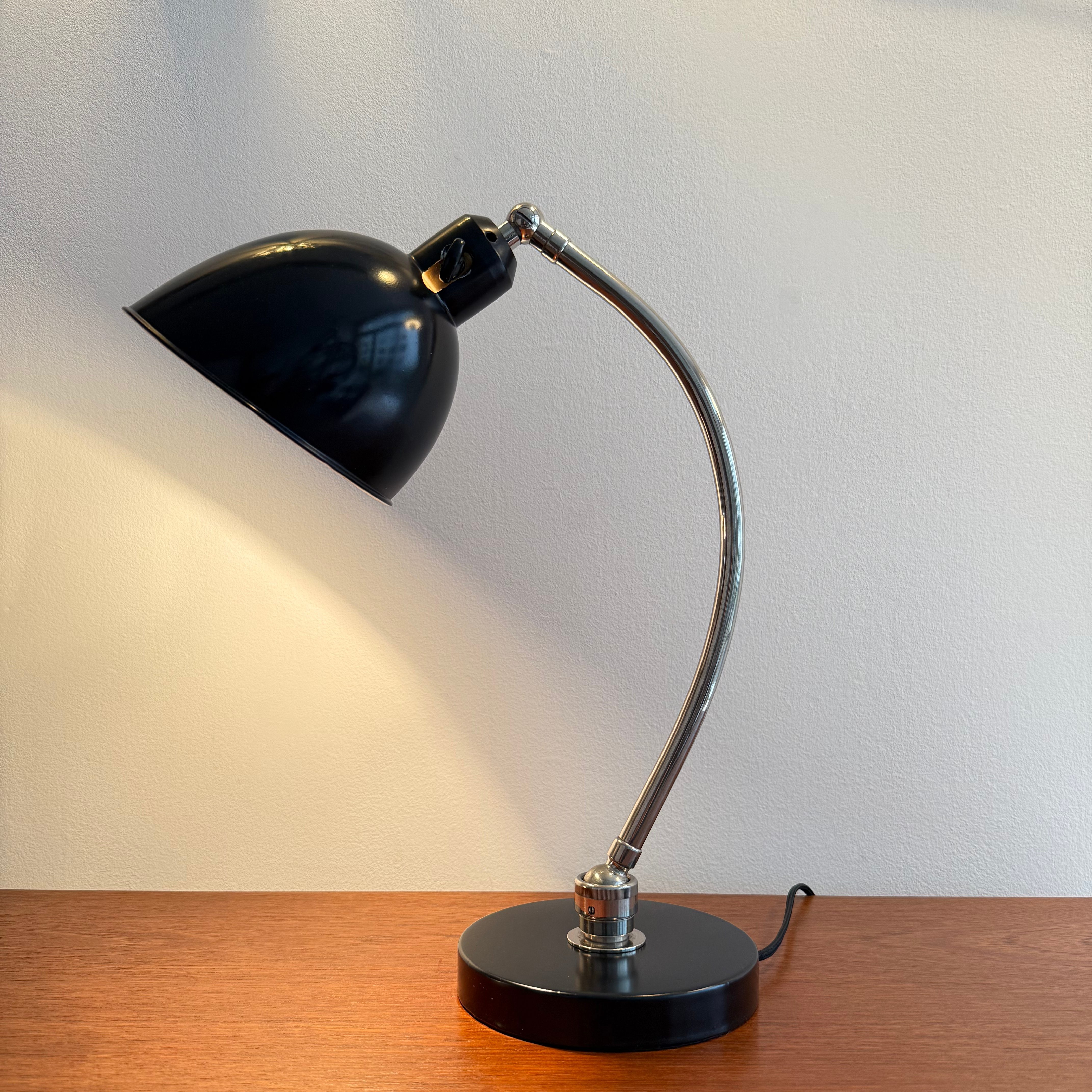 A mid-century modern black desk lamp with a curved metal arm and a circular base, illuminated and placed on a wooden surface against a white wall.