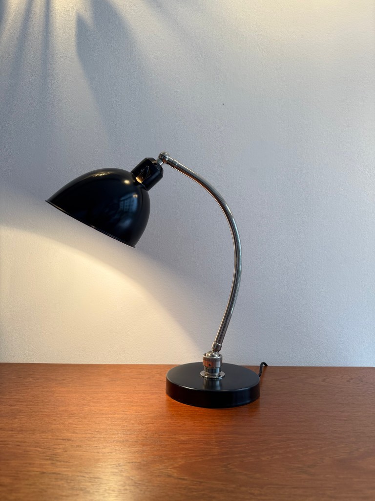 A black Bauhaus desk lamp with an adjustable curved arm and a circular base, placed on a wooden table against a plain wall.