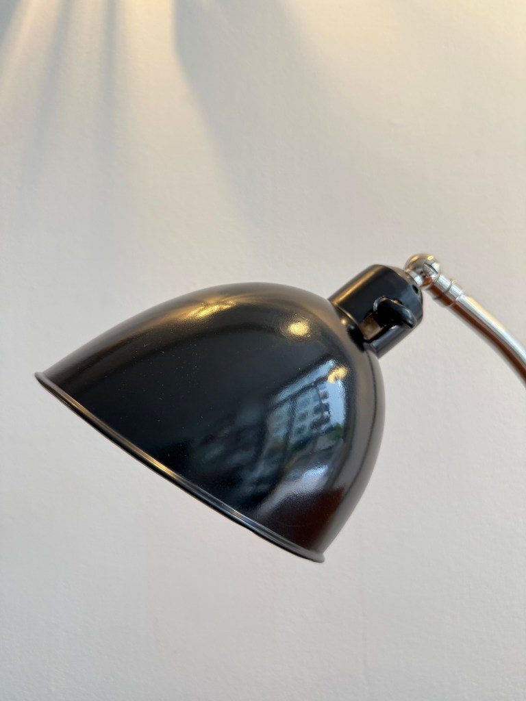 Close-up view of a Bauhaus desk lamp with a black shade and sleek metallic arm against a neutral background.