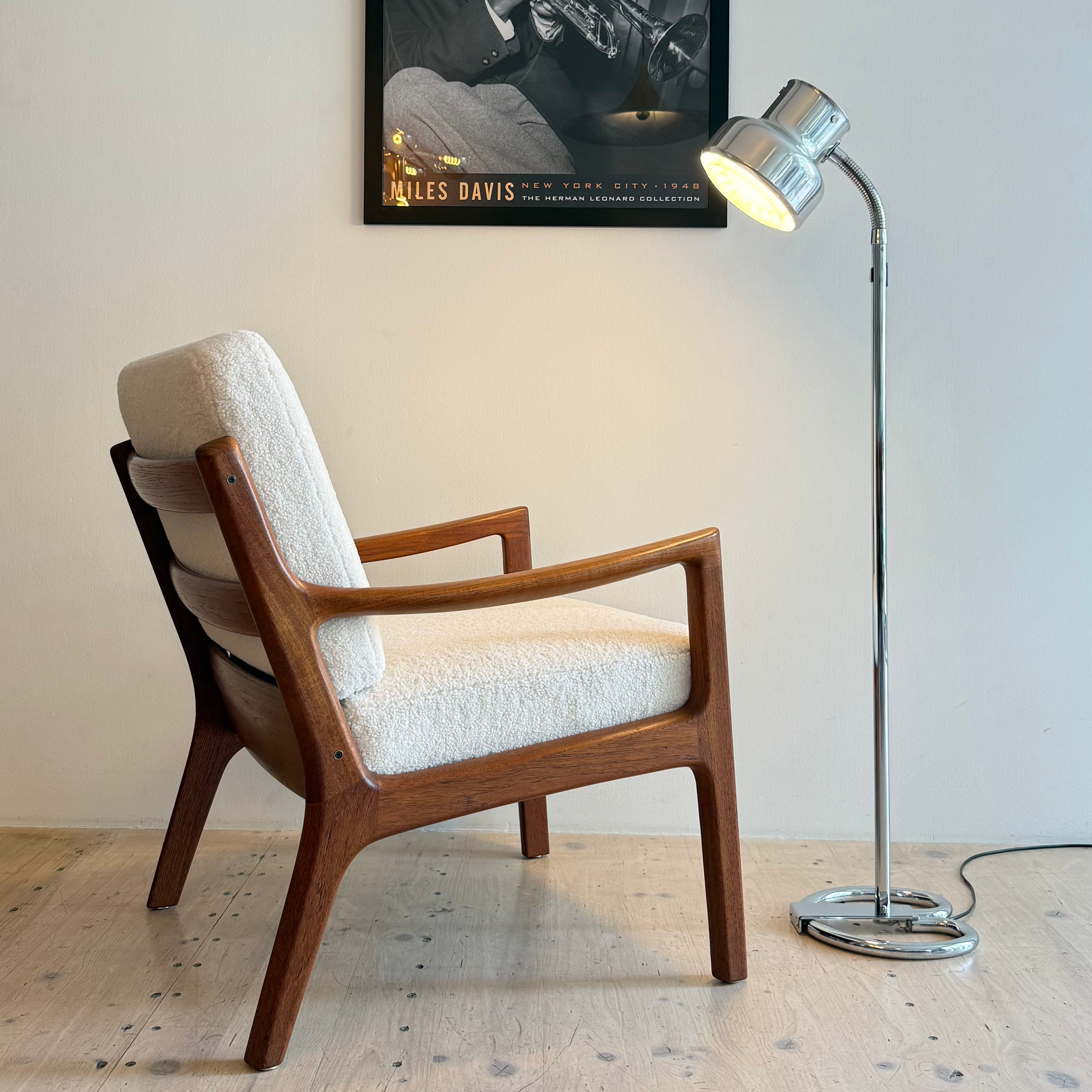 A stylish wooden chair with light upholstery next to a chrome floor lamp, set against a neutral wall featuring a framed black and white poster of Miles Davis playing the trumpet.