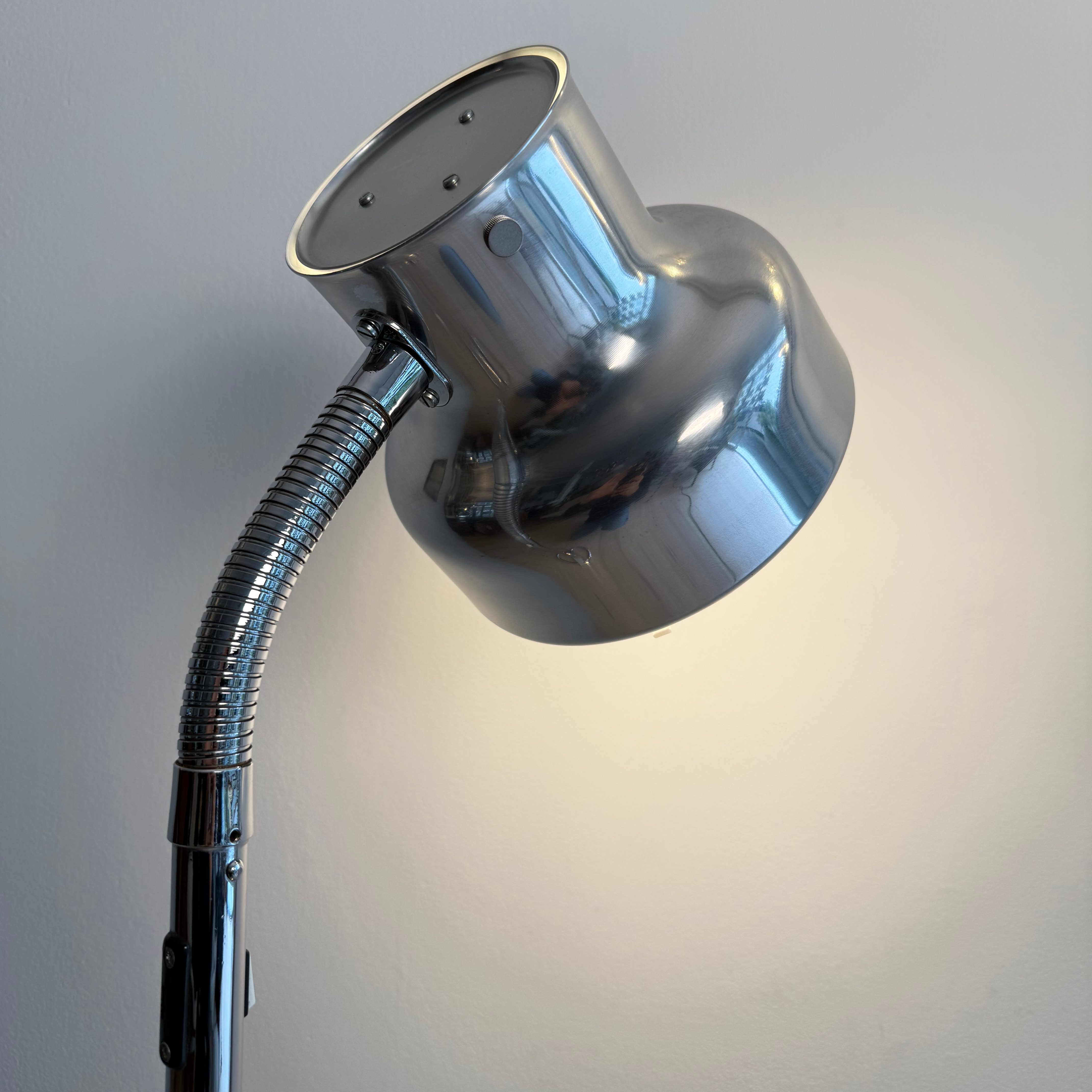 Close-up of a chrome floor lamp with a flexible neck and circular shade, emitting light against a plain wall.