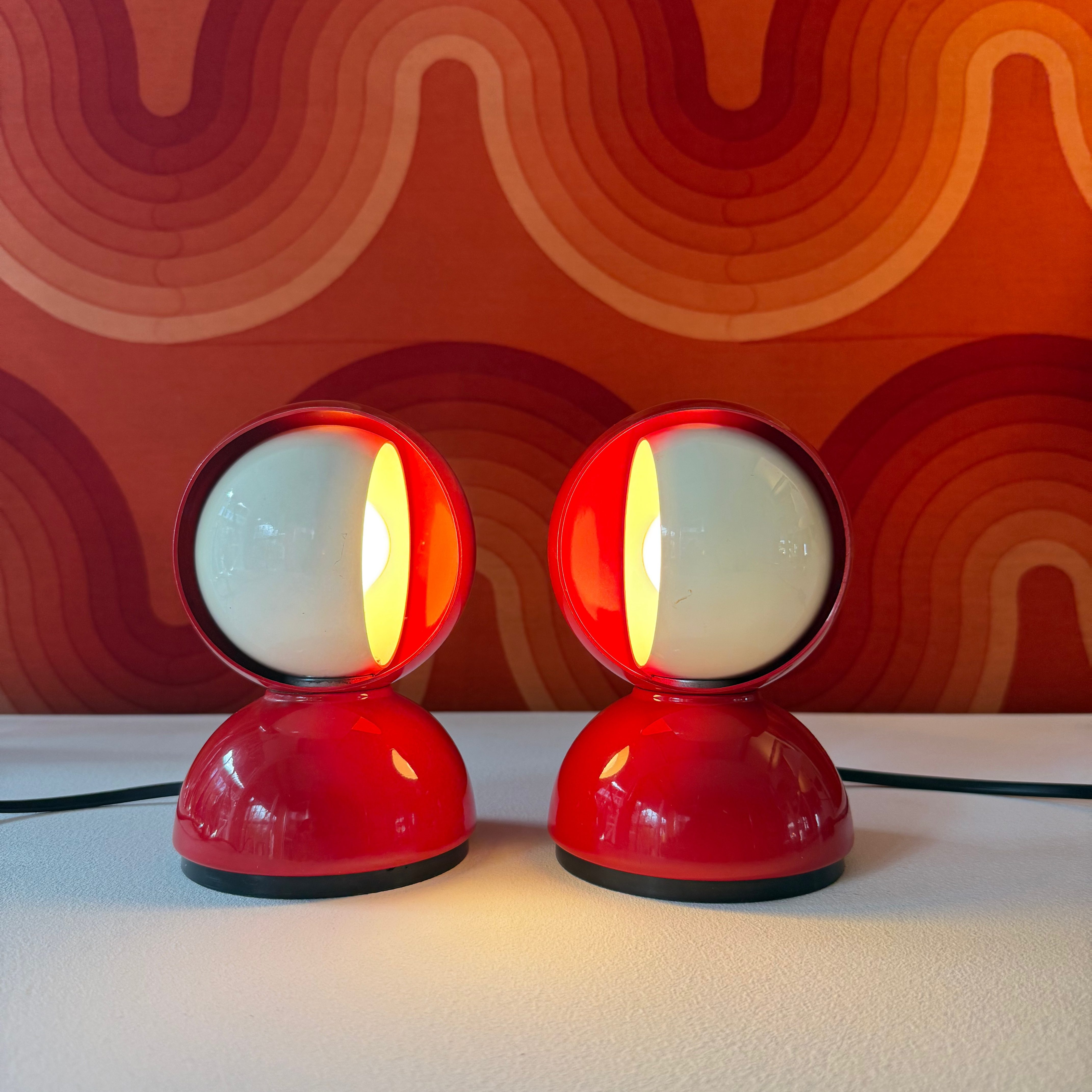 A pair of retro red mini desk lamps with spherical white shades, placed on a white surface, against a colorful wavy background.
