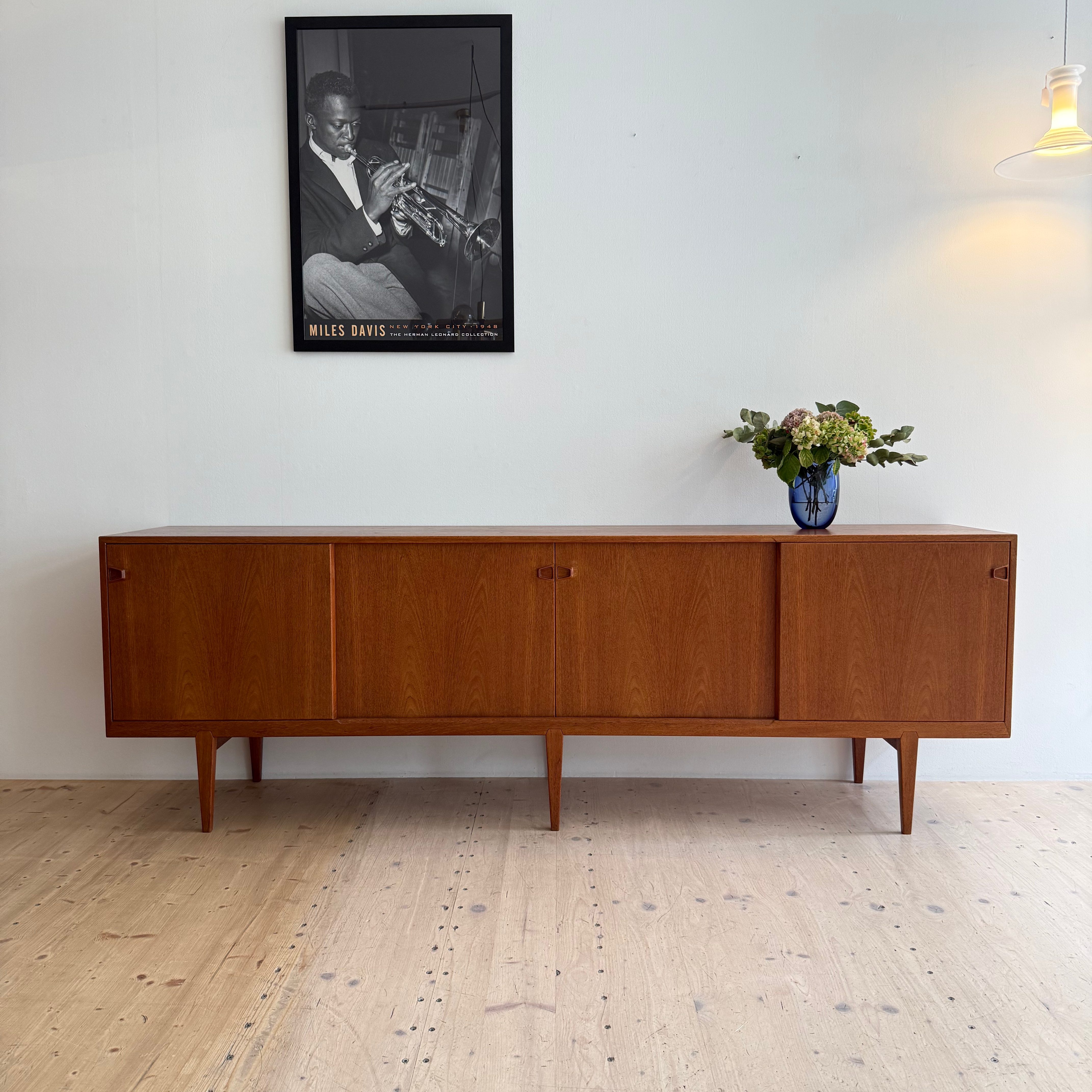 A wooden sideboard with sliding doors, placed against a light-colored wall. Above it, there is a framed black-and-white photograph of Miles Davis, and a vase with fresh flowers is on the sideboard.