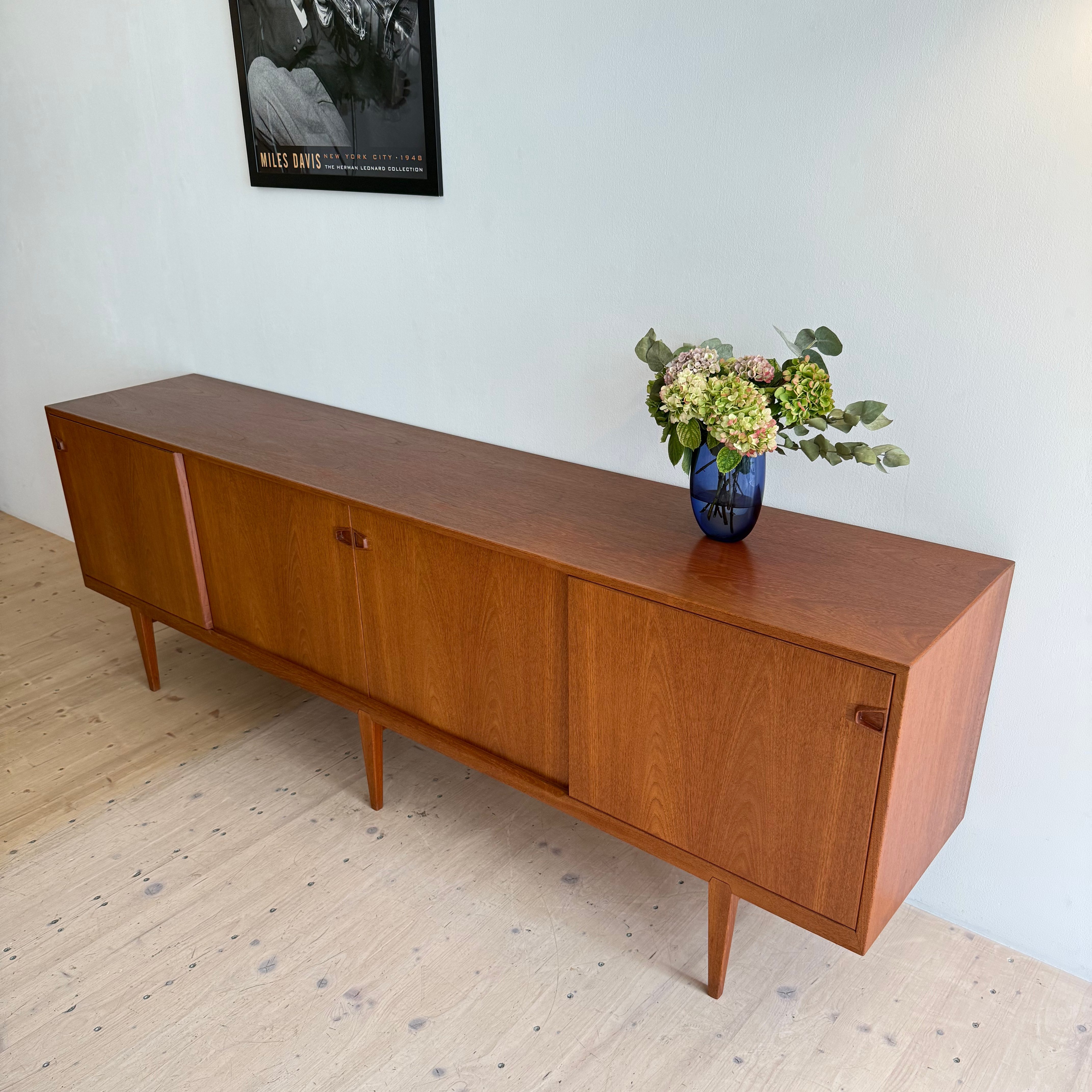 A sleek Danish teak sliding door sideboard with two sliding doors, displayed against a light-colored wall. A vase of flowers sits on top, and a black-and-white poster is visible in the background.