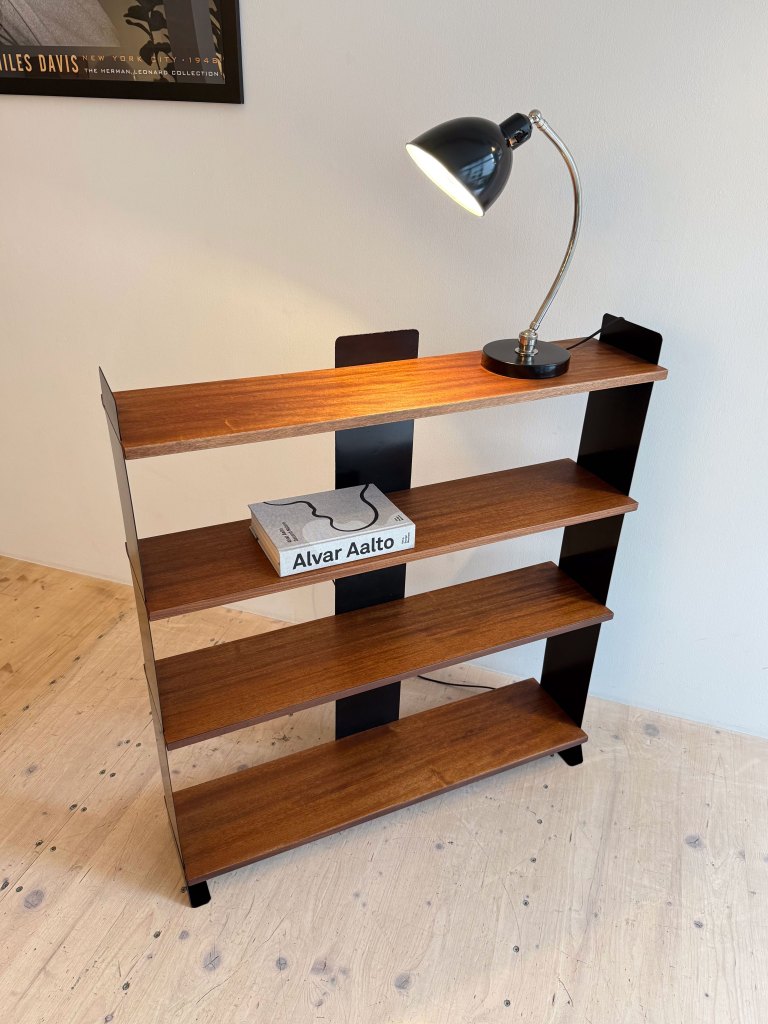 A wooden bookshelf with four shelves, featuring a black lamp on the upper shelf and a book titled 'Alvar Aalto' on the middle shelf.