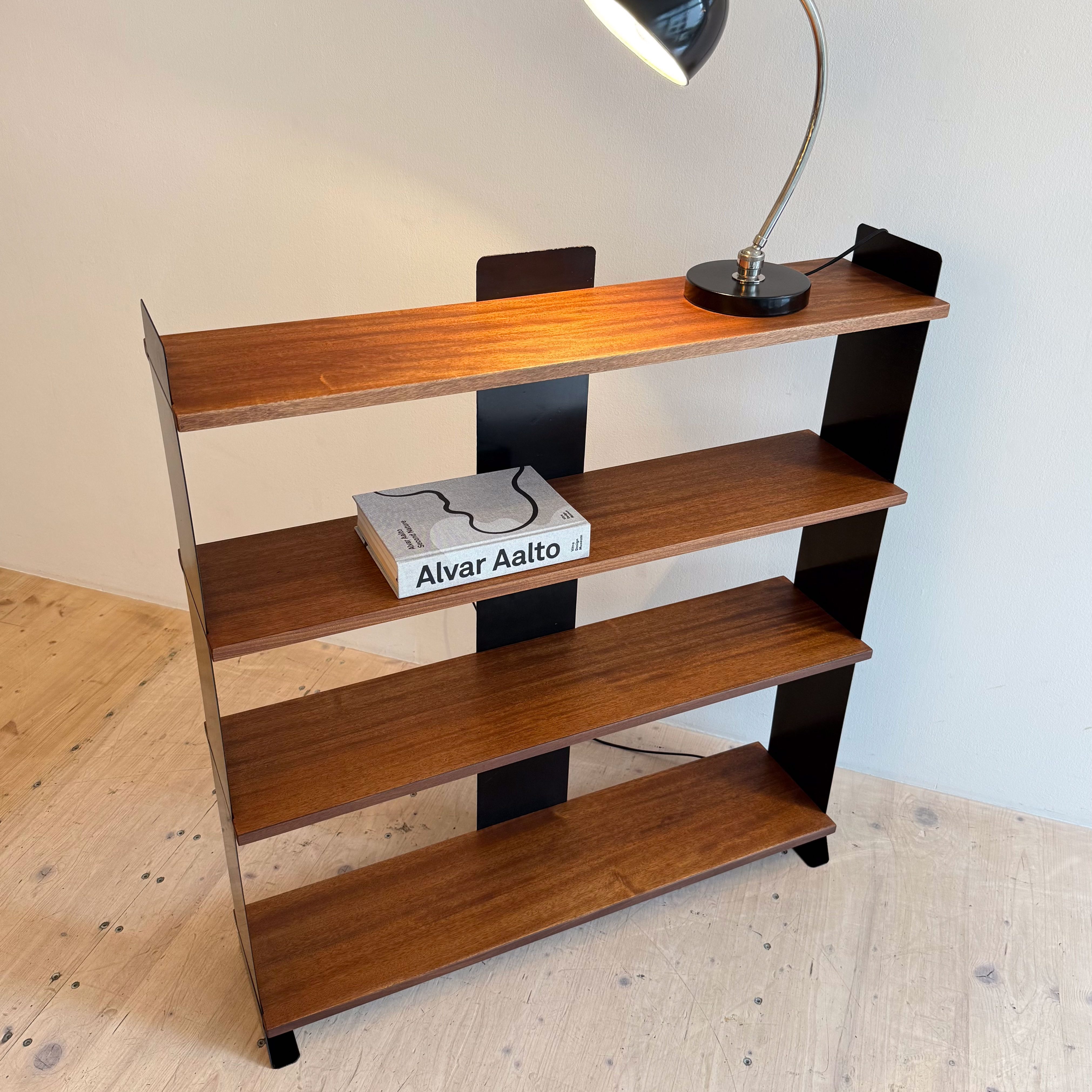 A modern wood shelving unit with four shelves, featuring a sleek black lamp and a book by Alvar Aalto on the top shelf.