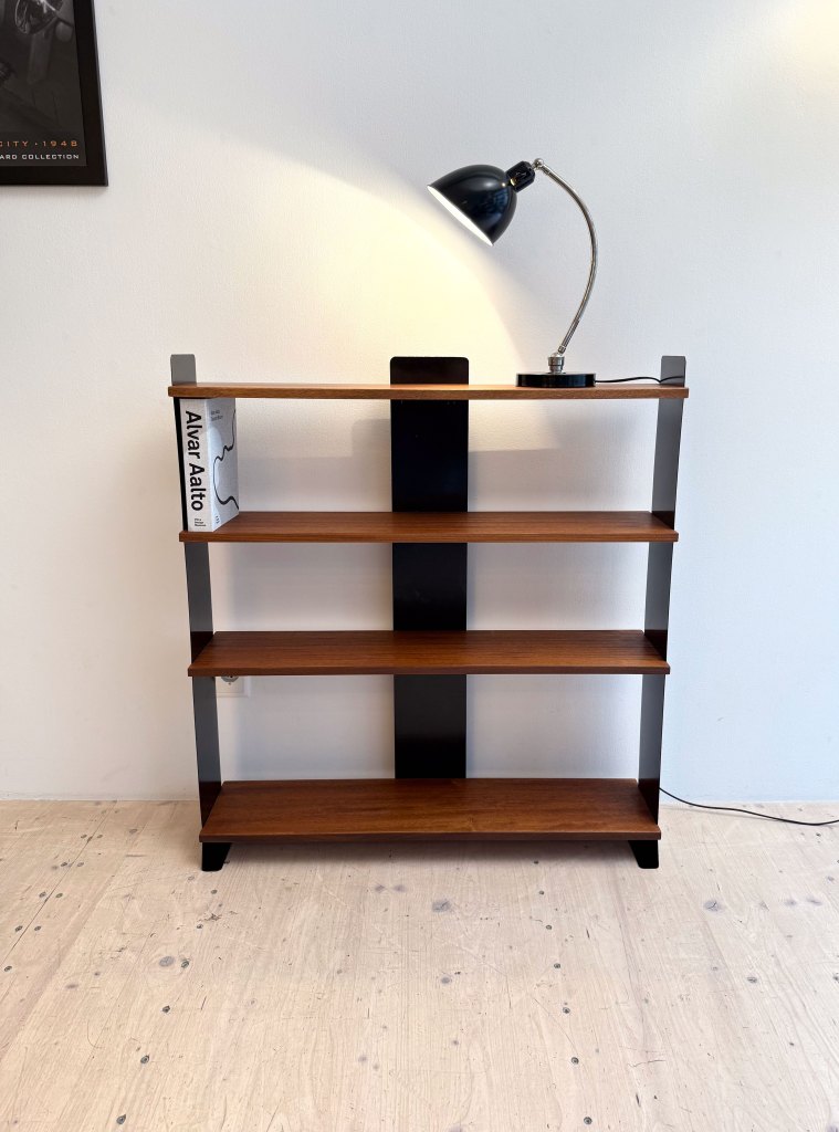 A modern bookshelf with three wooden shelves, a black lamp on top, and a notable book titled 'Alvar Aalto'.
