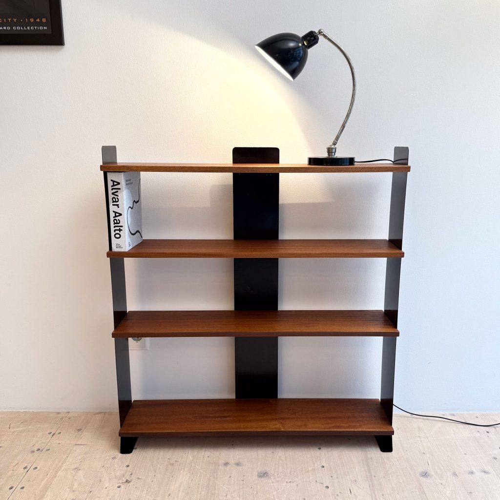 A mid-century modern bookshelf with wooden shelves and a black lamp, featuring a book titled 'Alvar Aalto' on the top shelf, placed against a plain wall.