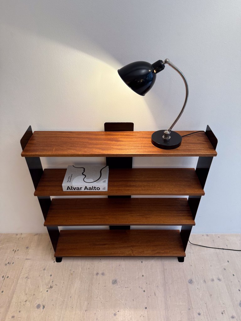 A modern wooden shelf with black accents, featuring three open shelves and a black desk lamp illuminating the top surface. A book titled 'Alvar Aalto' is placed on one of the shelves.