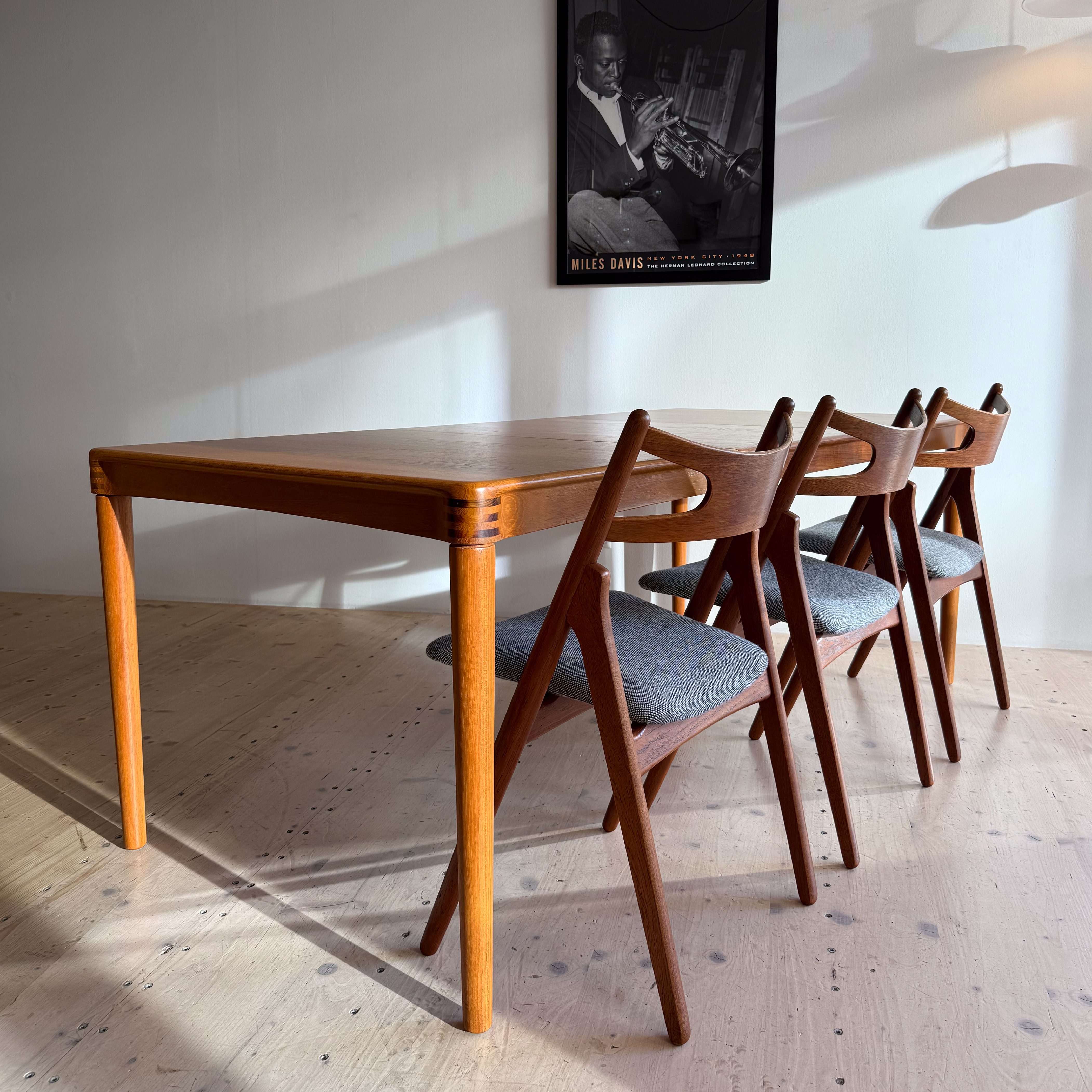 A wooden dining table surrounded by four matching chairs, with a black and white poster of Miles Davis on the wall.