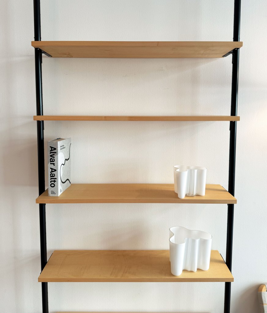 A modern shelving unit with wooden shelves and black metal frame, featuring a book titled 'Alvar Aalto' and two white vases on the middle shelves.