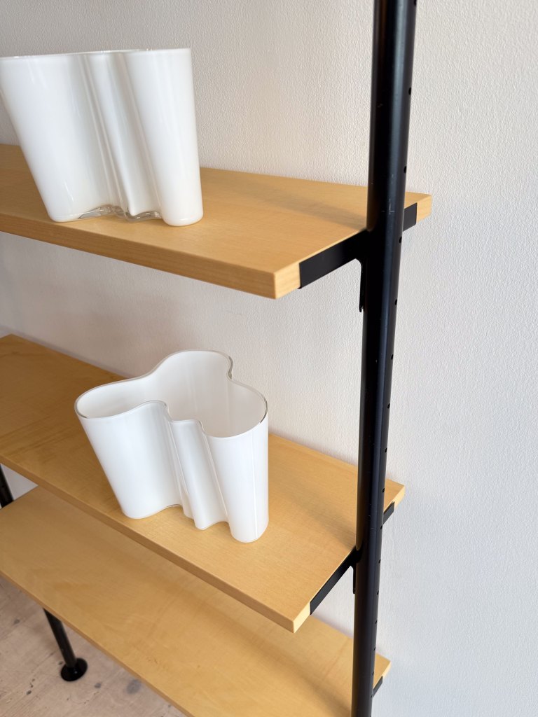 A close-up view of a wooden shelf with two unique white vases. One vase is shaped like a wave and the other has a more conventional form. The background is a simple white wall.