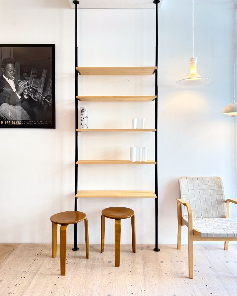 A modern interior setting featuring a tall, minimalist wooden shelf with black supports against a white wall. Below the shelf, there are two round wooden stools and a stylish woven chair. A poster of a musician hangs on the wall, and soft lighting illuminates the space.