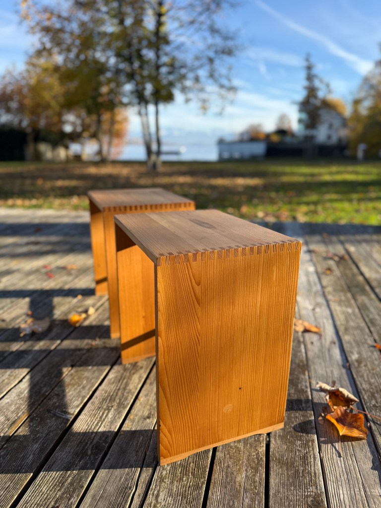 Two wooden stools placed on a wooden deck, surrounded by autumn foliage and a calm body of water in the background.