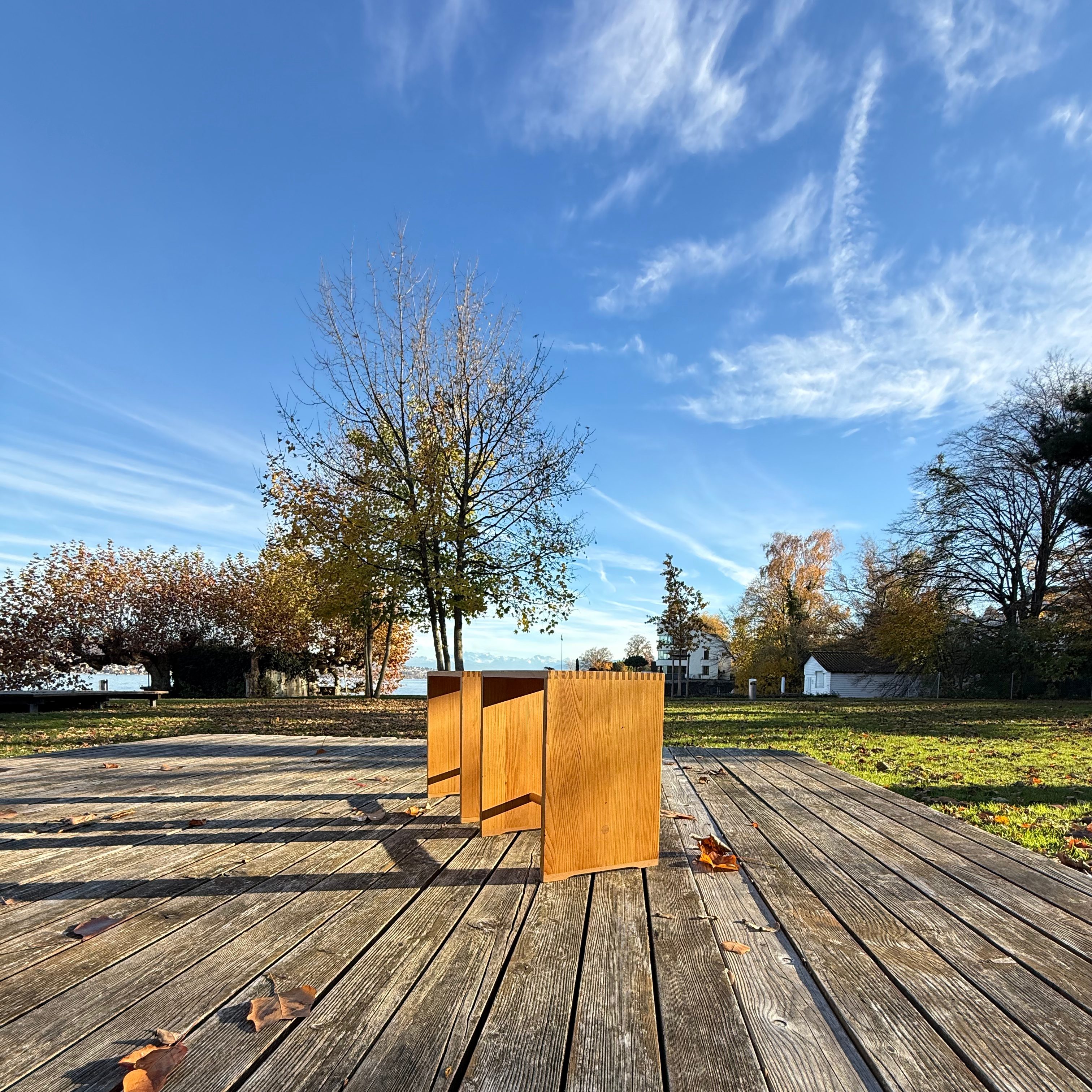 A wooden stool placed on a wooden deck, surrounded by autumn leaves and trees under a clear blue sky.
