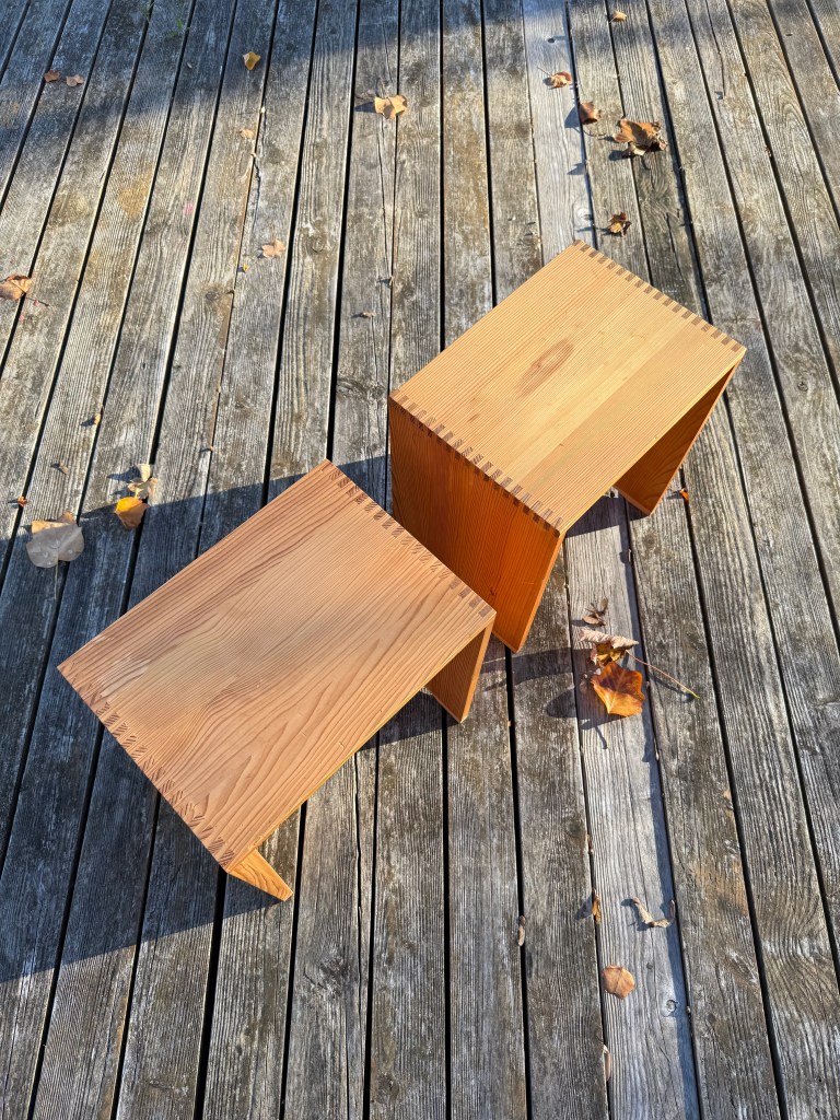 Two wooden stools with a simple design on a wooden deck, adorned with a few scattered leaves.
