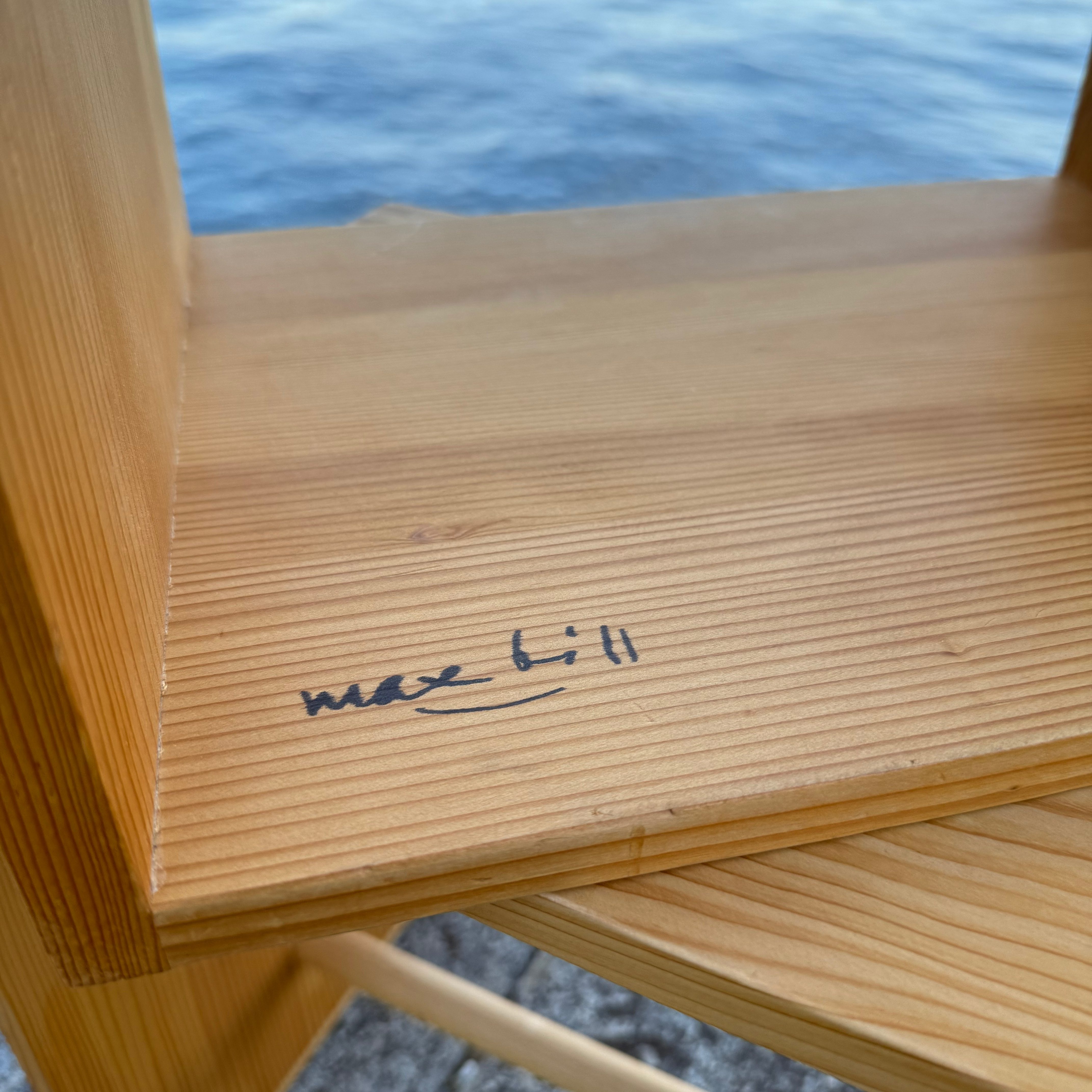 Close-up of a wooden shelf displaying the signature 'max bill', with water in the background.