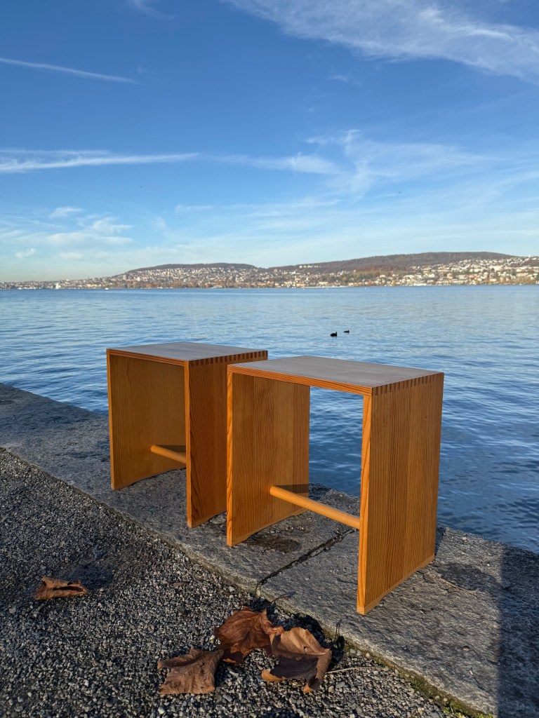 Two wooden stools placed on a stone ledge by a body of water, with a scenic view of a city and mountains in the background under a clear blue sky.
