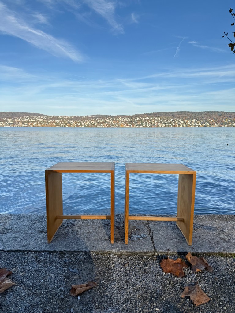 Two wooden side tables positioned next to a calm lake, with a scenic background of a shoreline and blue sky.