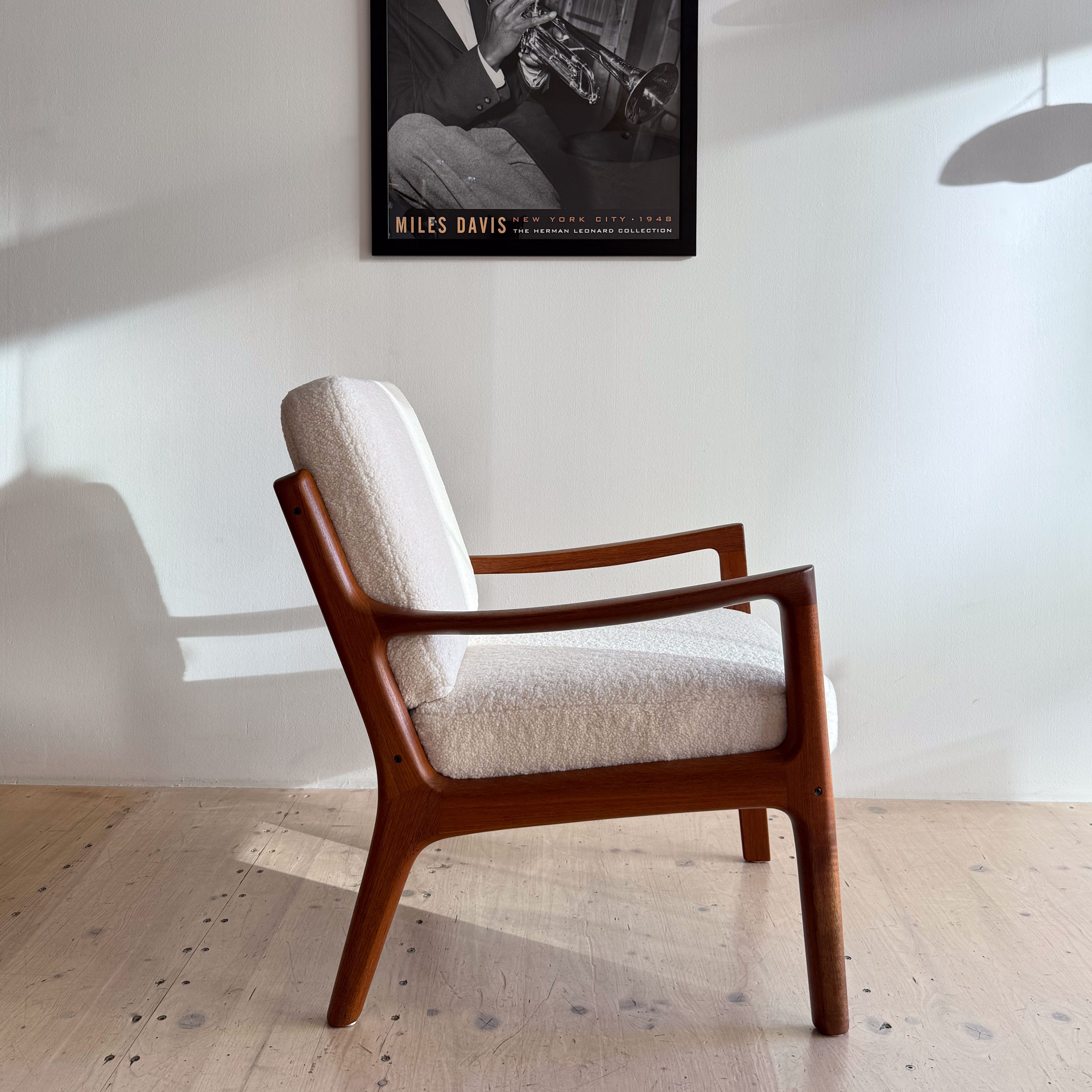 A mid-century modern lounge chair with a wooden frame and light fabric upholstery, positioned next to a wall featuring a black and white poster of a jazz musician playing the trumpet.