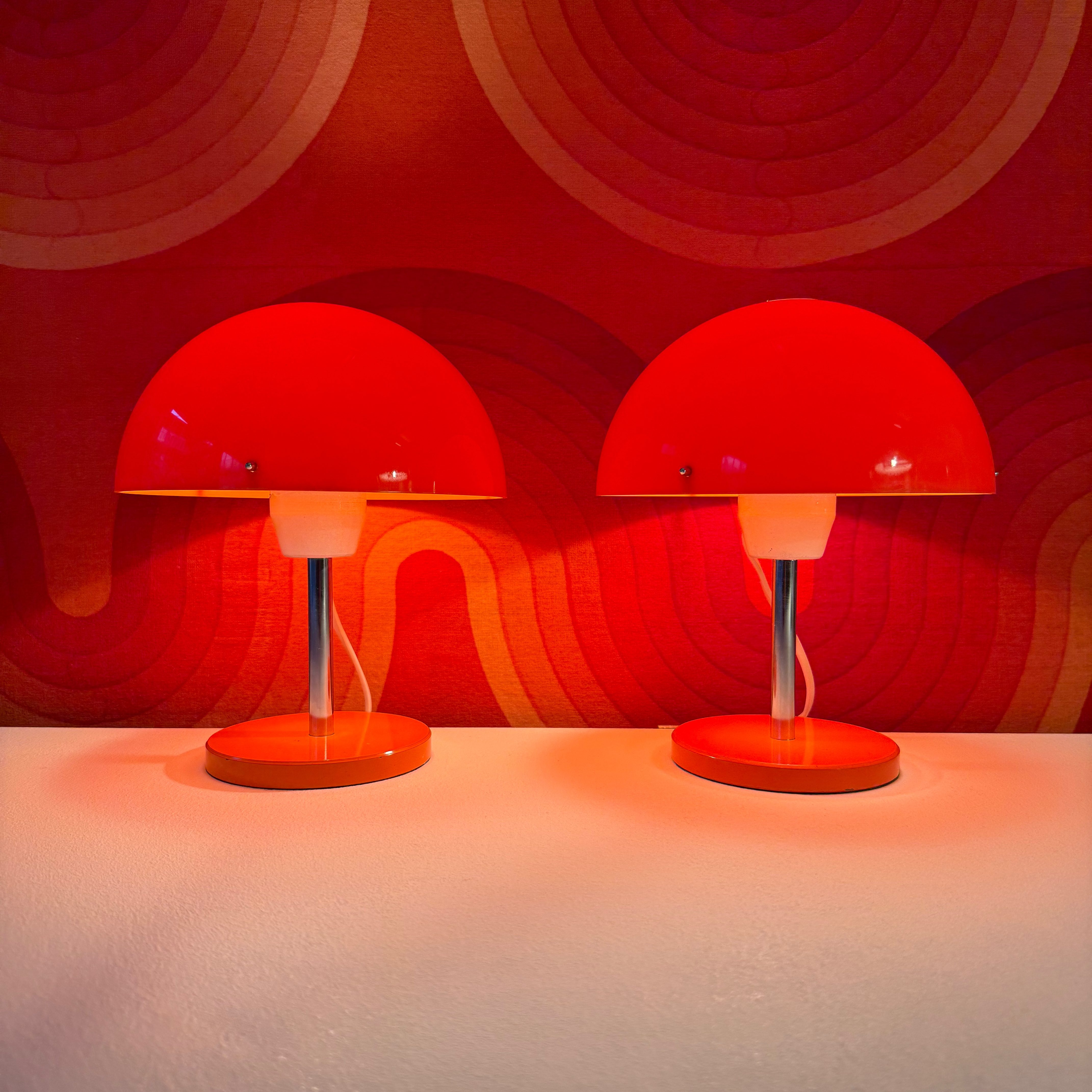 Two vibrant red table lamps with circular shades and a glossy base, set against a warm, patterned background.