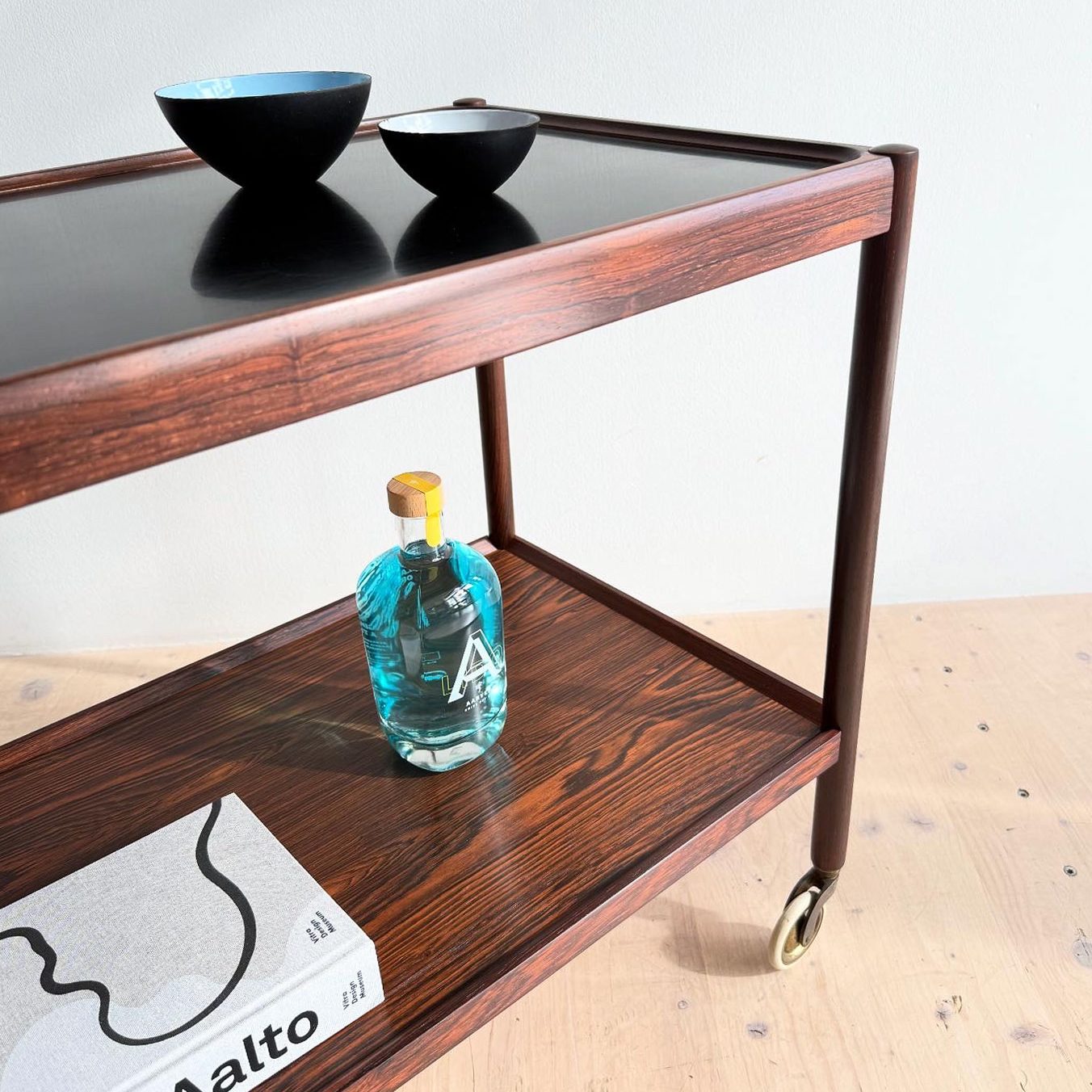 Teak serving cart with a black top, two decorative bowls, a bottle, and a book displayed on it.